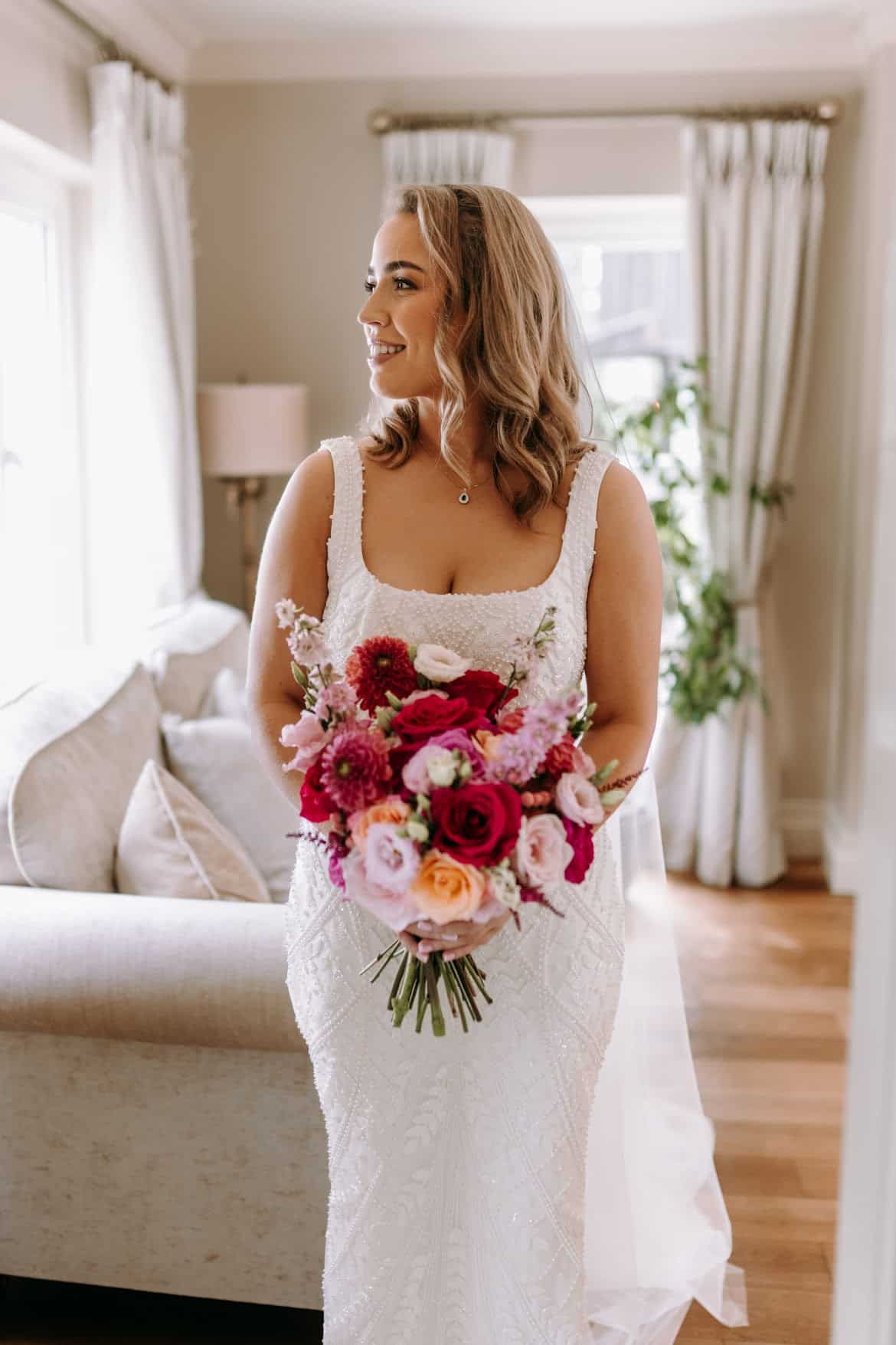 Bride in white dress holding a vibrant bouquet, standing in a bright, elegantly decorated room.