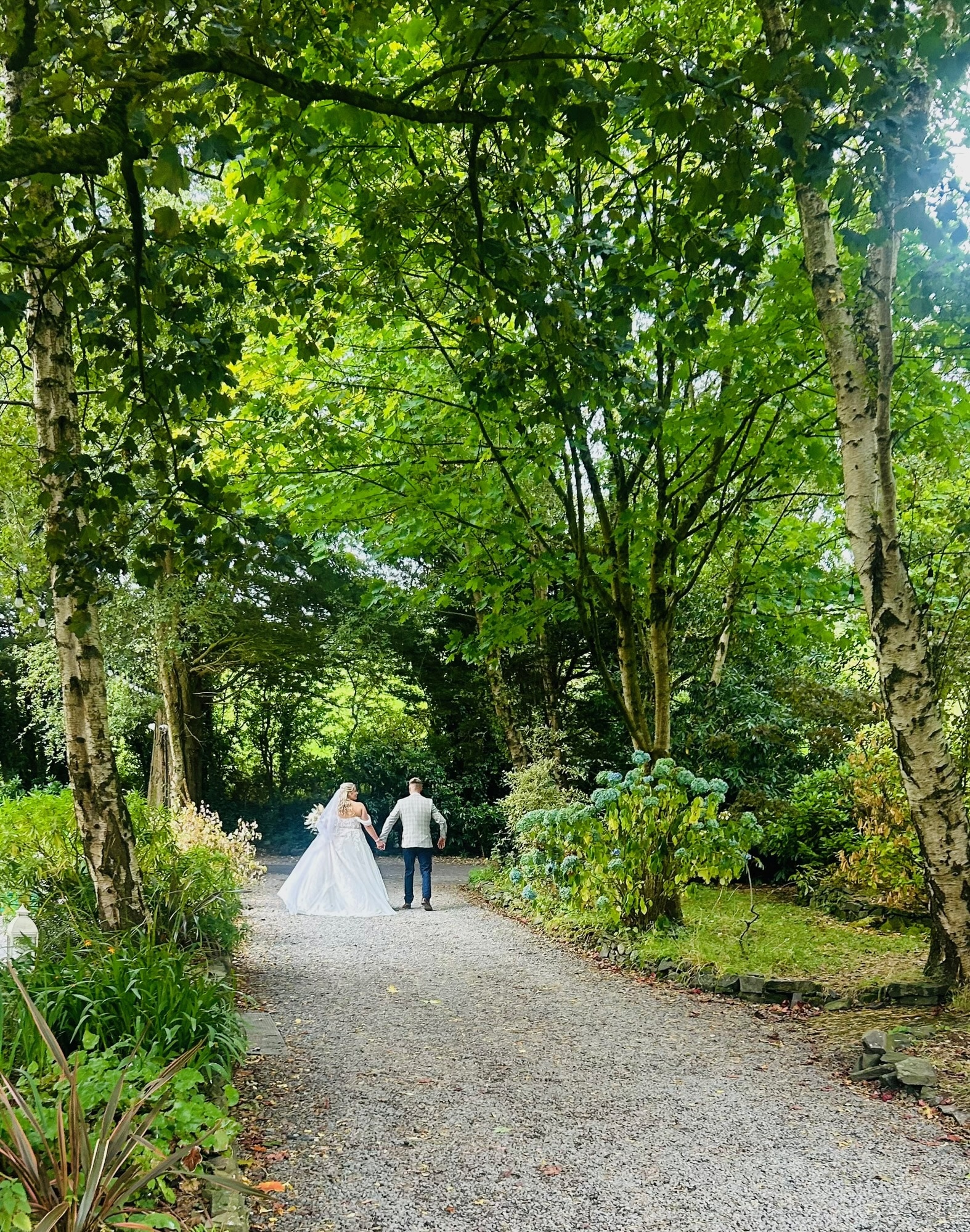 Bride and groom walking hand in hand down a tree-lined gravel path in a lush garden.