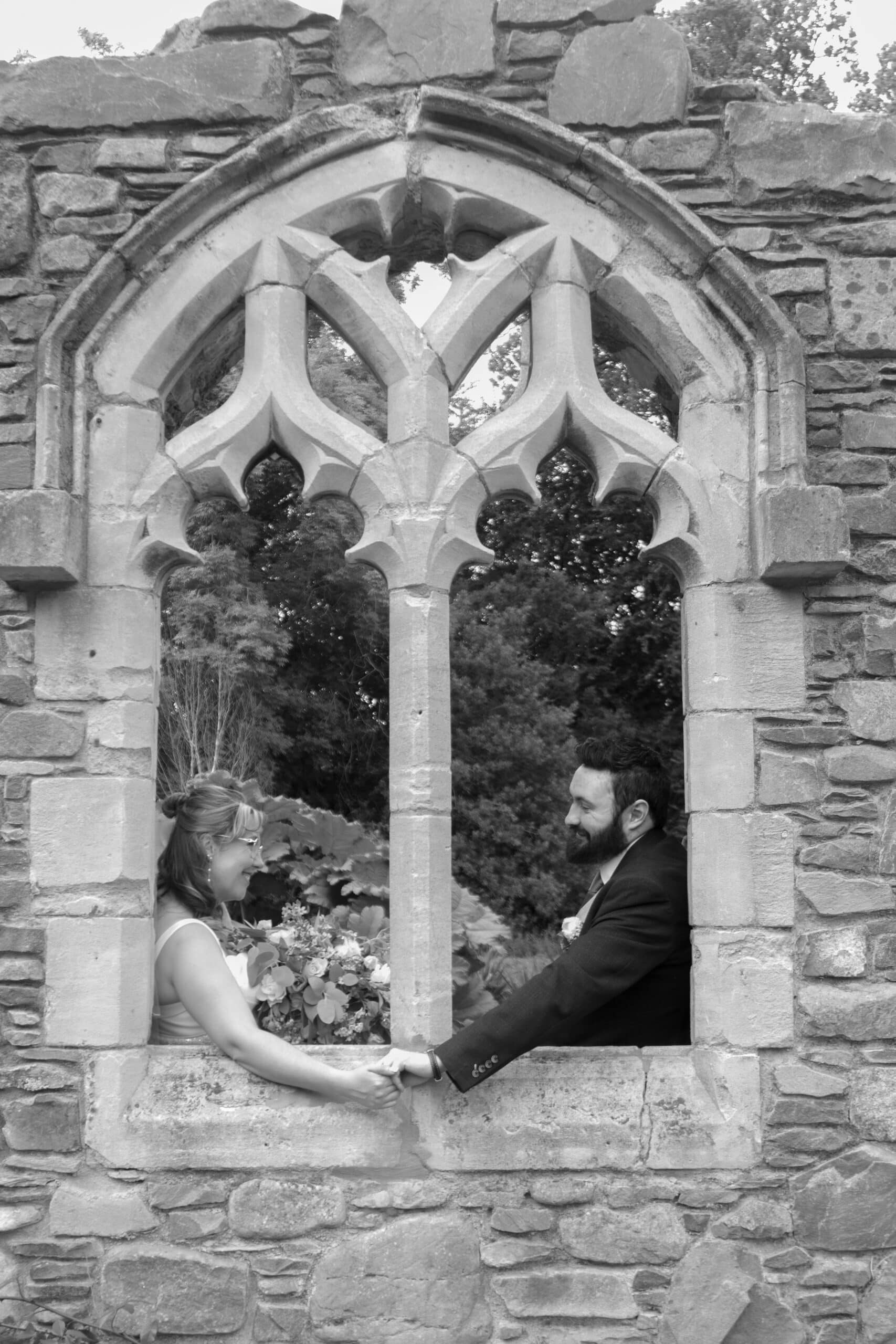Bride and groom holding hands through a church ruin window, black and white photo.
