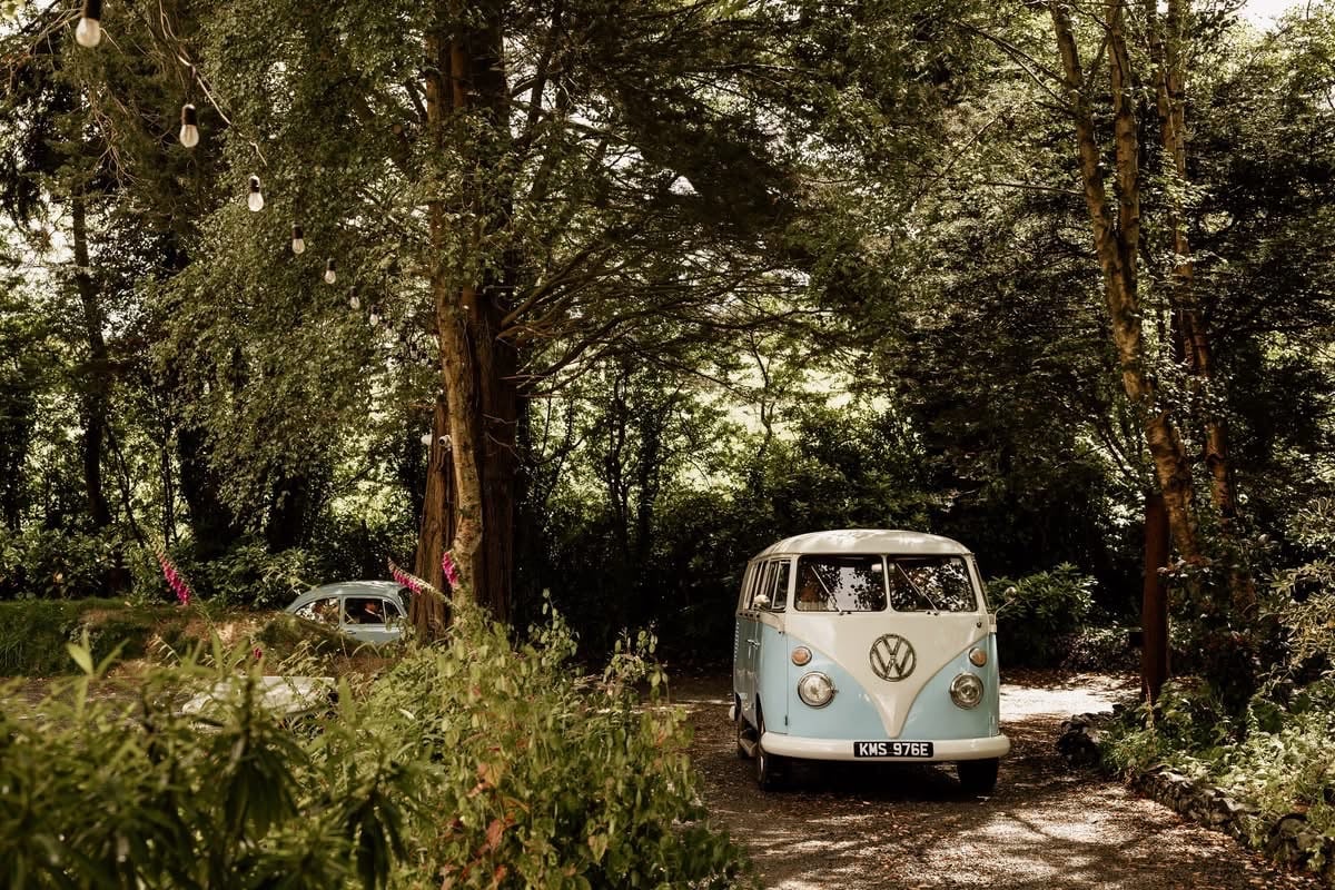 Vintage blue VW van parked on a wooded path surrounded by lush greenery and outdoor string lights.