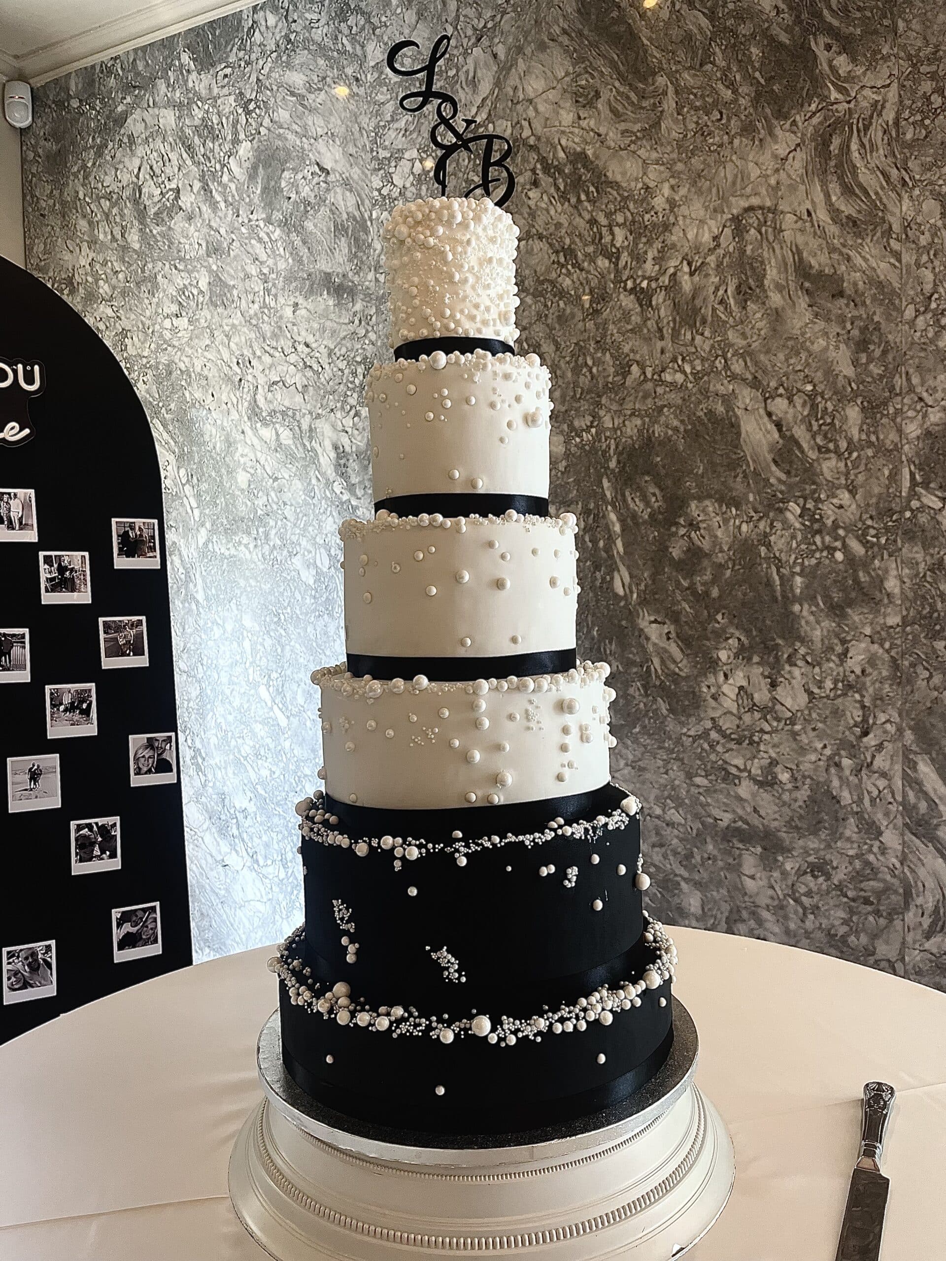 Elegant six-tier black and white wedding cake with pearl decorations and initial topper on a marble background.