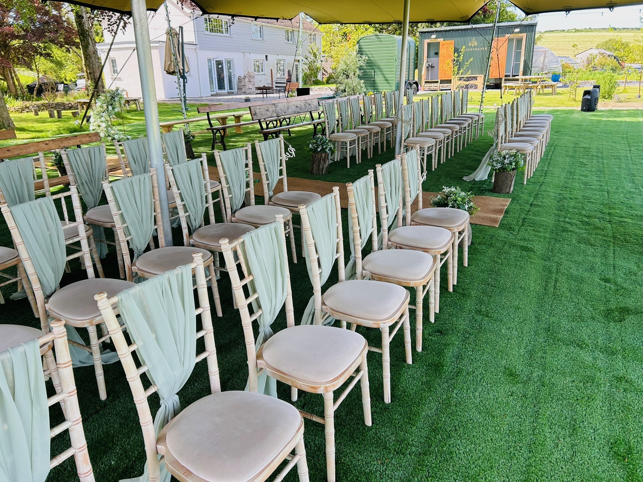 Outdoor wedding seating with white chairs draped in light fabric on green grass under a canopy.