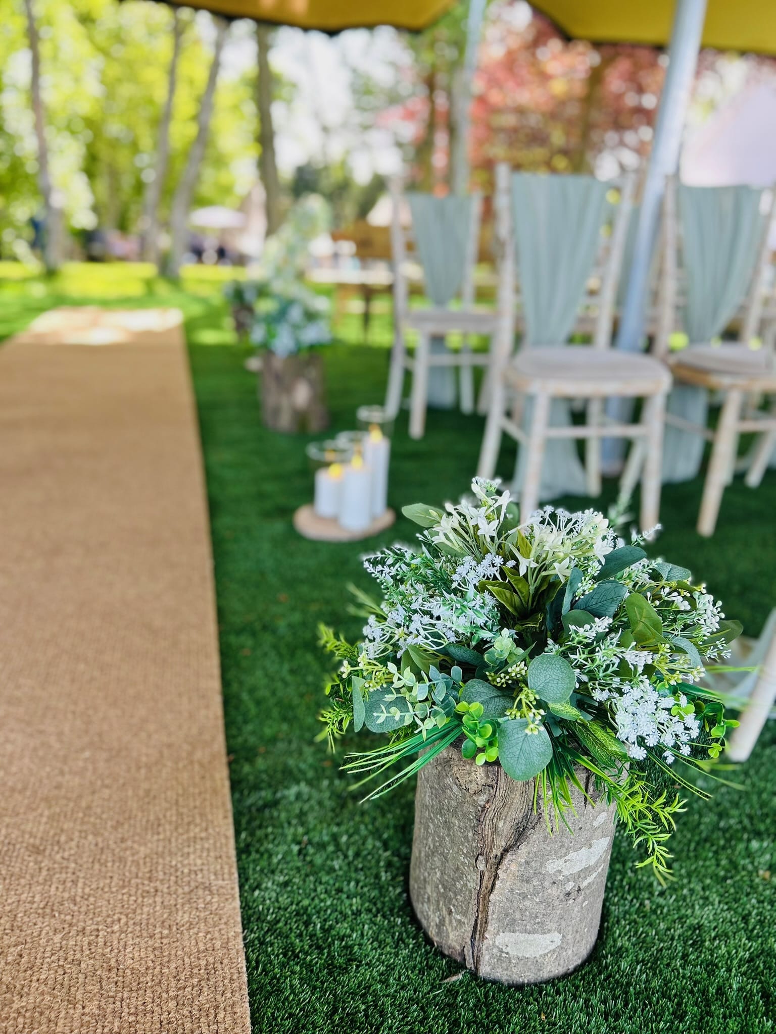Outdoor wedding aisle with wooden log floral arrangement and chairs on green grass.