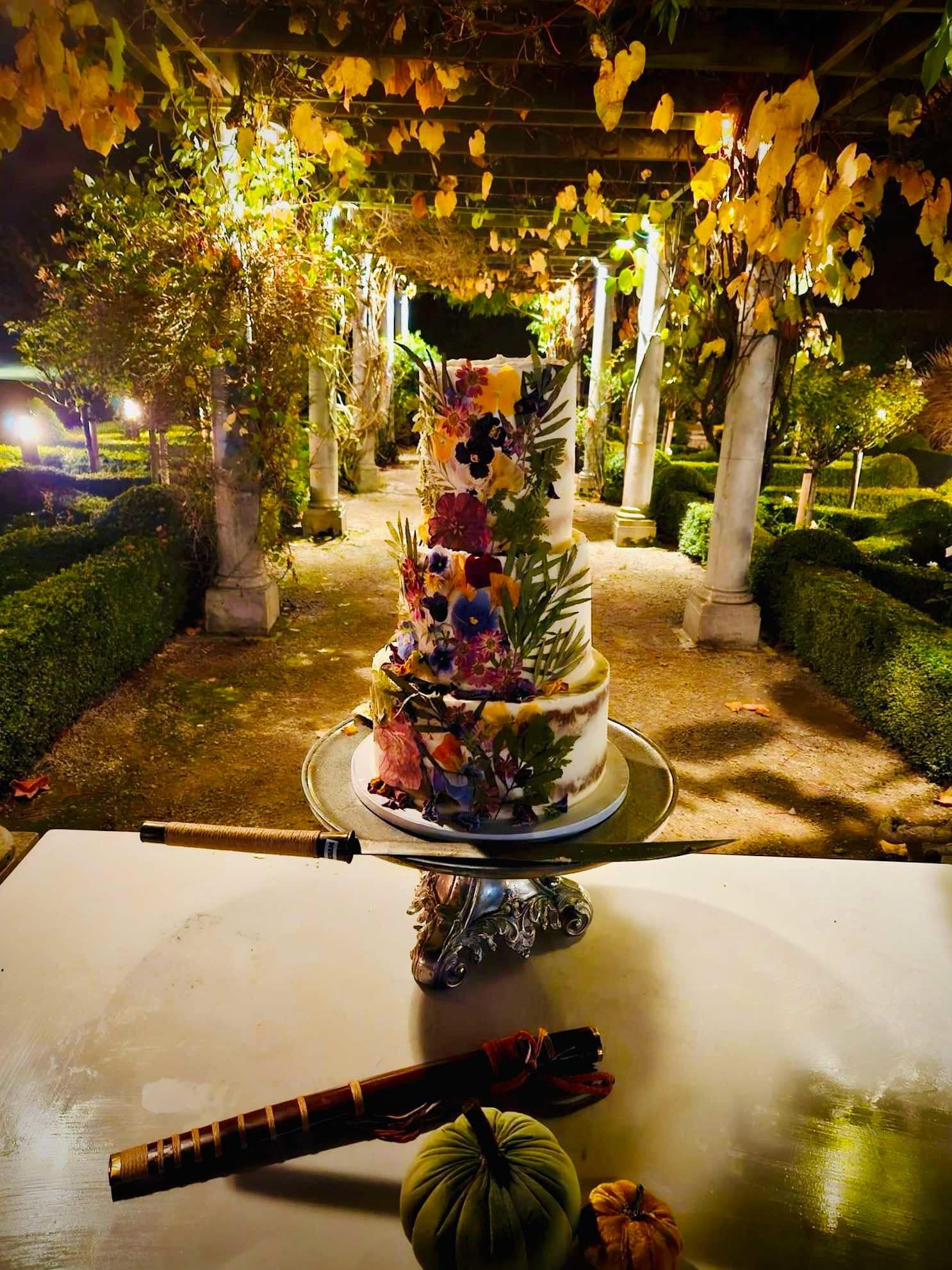 Three-tiered floral cake under a pergola with ivy-covered columns at night, with decorative items on the table.
