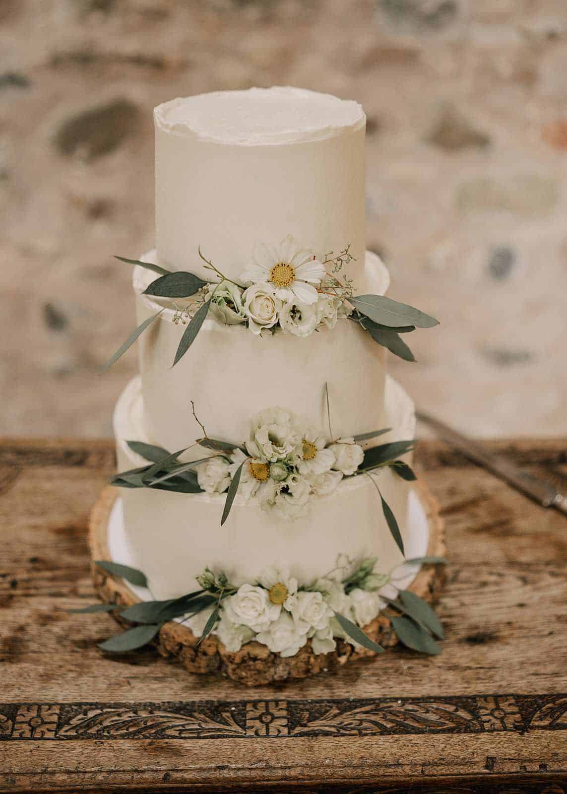 Three-tier white wedding cake with floral decorations and green leaves on each tier, set on a rustic wooden table.