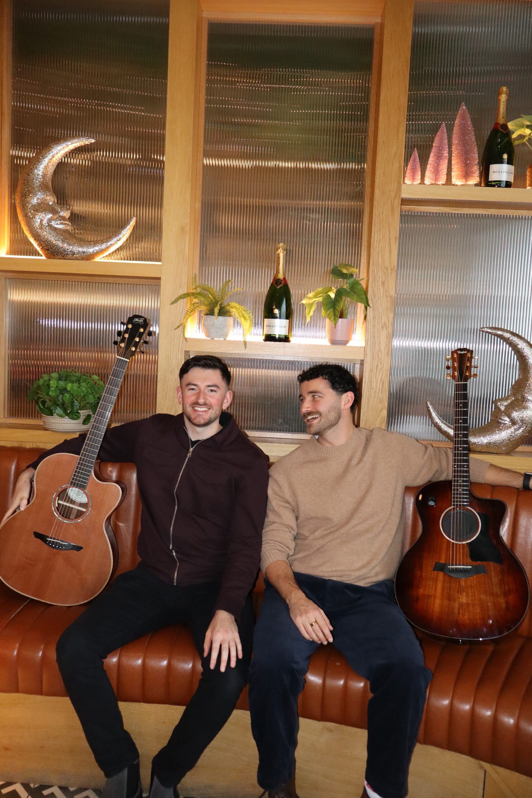Two men sitting on a leather couch with guitars, surrounded by decorative shelves with plants and champagne bottles.