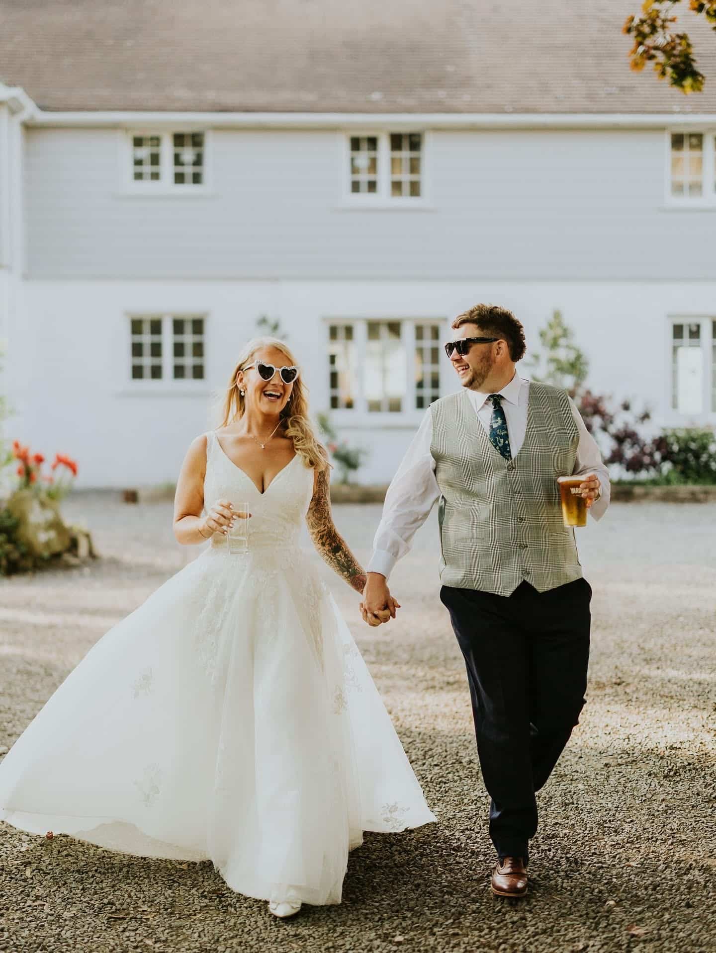 Bride and groom in sunglasses walking hand in hand outside a house, smiling and holding drinks.