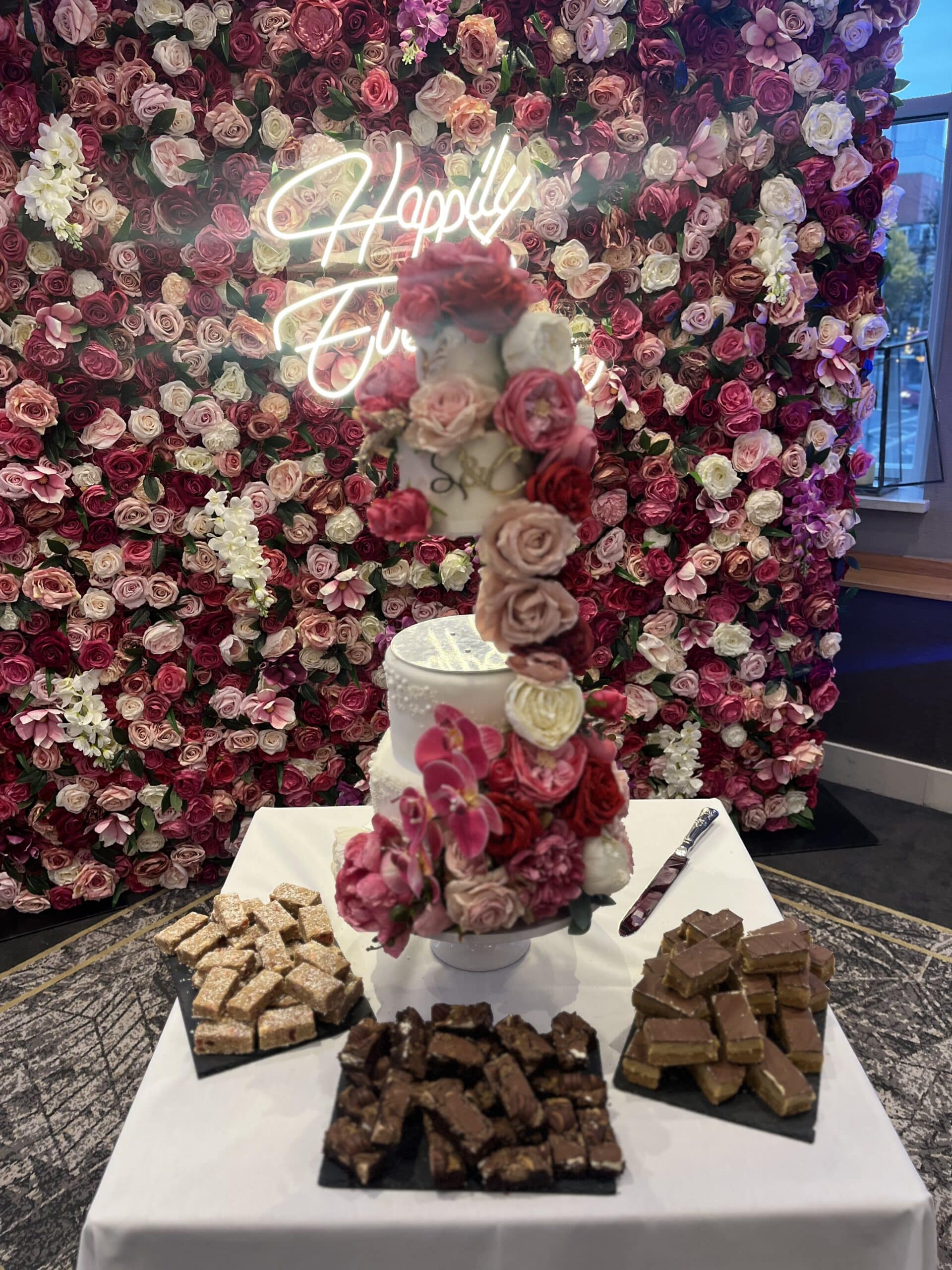 Floral wedding cake and treats on a white tablecloth, set against a vibrant rose wall with a neon "Happily Ever After" sign.