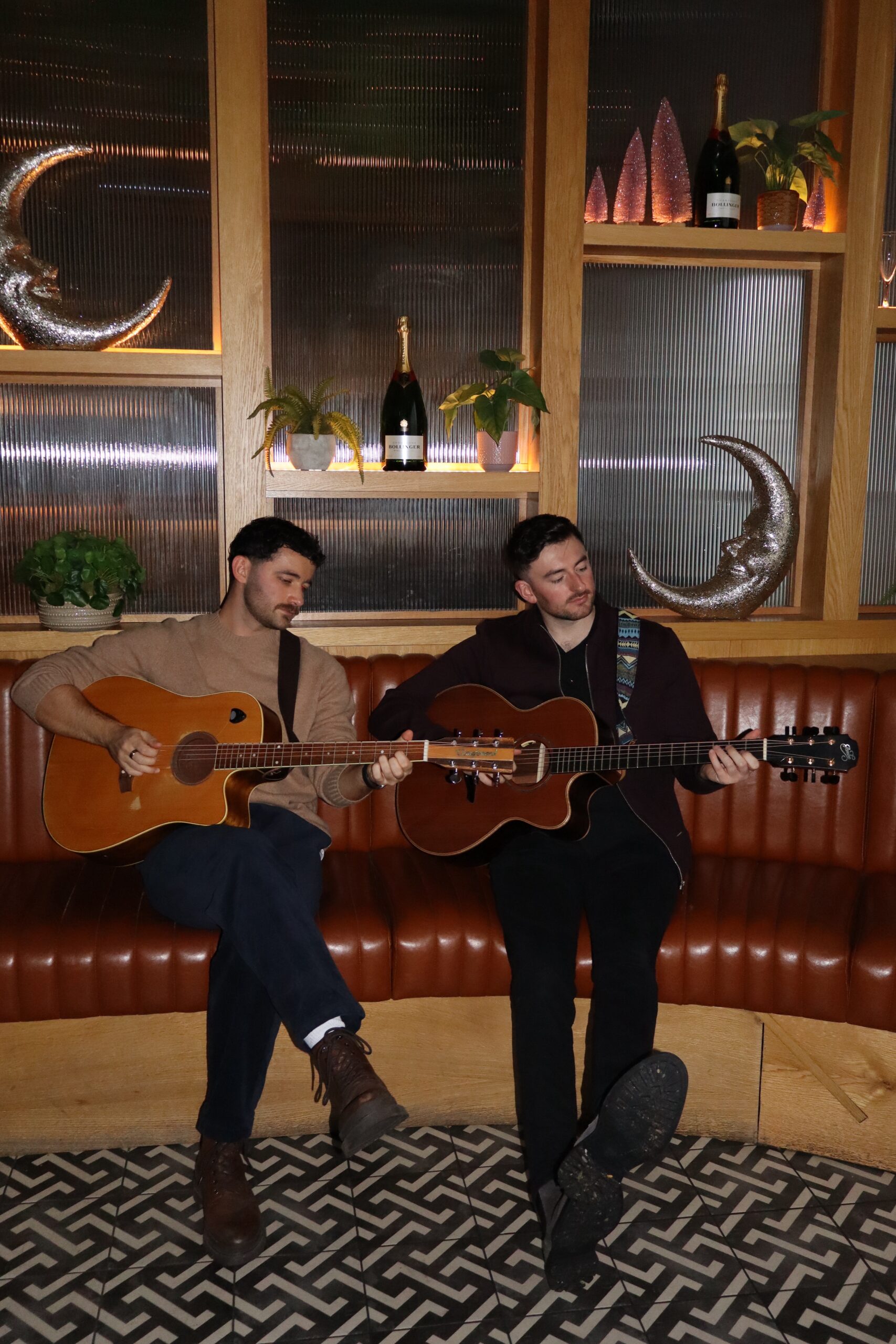 Two men playing acoustic guitars on a leather couch in a stylish room with decorative plants and bottles on shelves.