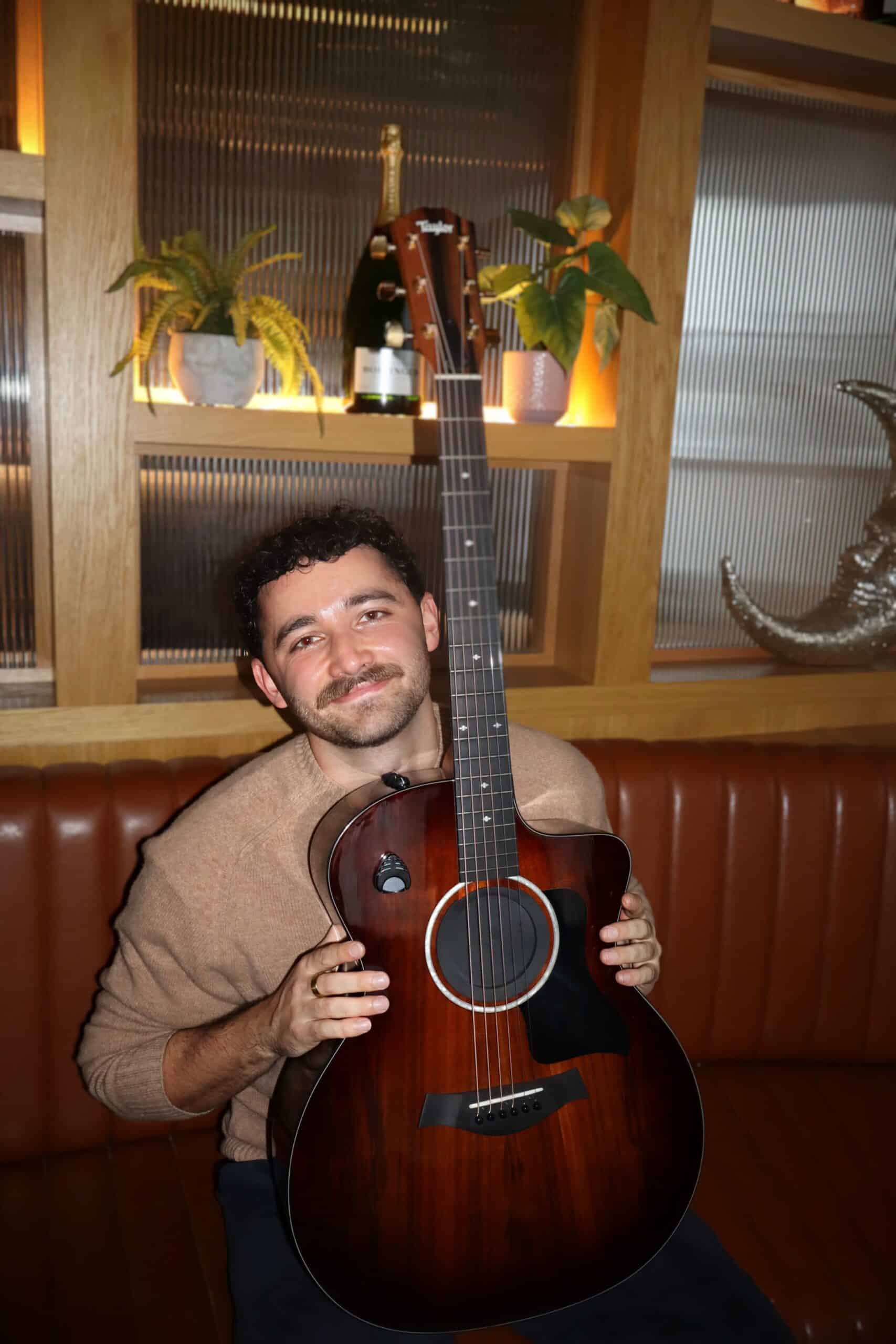 Man smiling while holding an acoustic guitar in a cozy room with plants and decorative items on shelves.