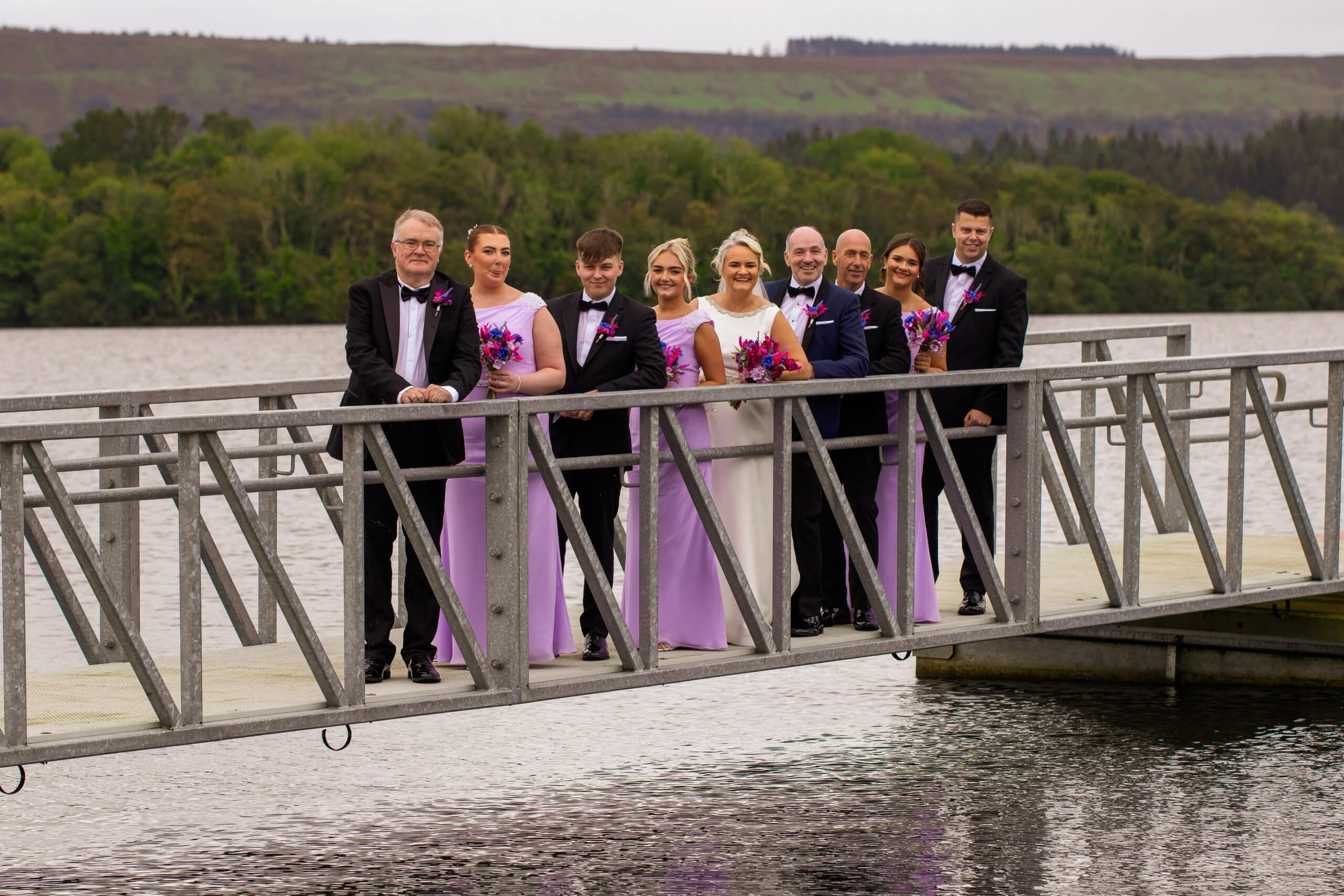 Wedding party on a dock, with bridesmaids in lavender dresses and groomsmen in black suits, holding bouquets.