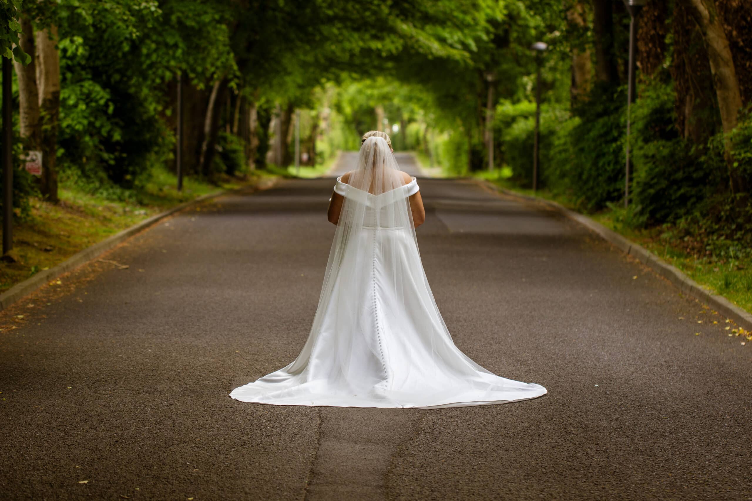 Bride in white gown standing on a tree-lined road, back view.