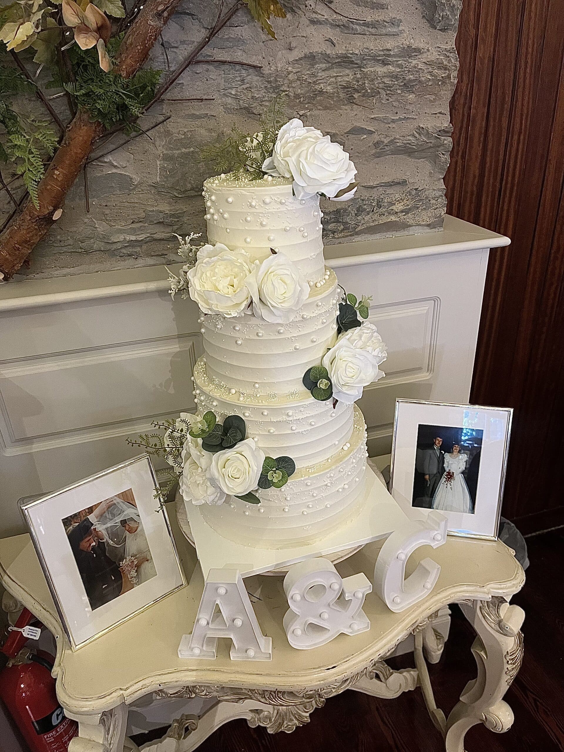 Elegant white wedding cake with floral decorations on a vintage table, accompanied by framed wedding photos and initials A&C.