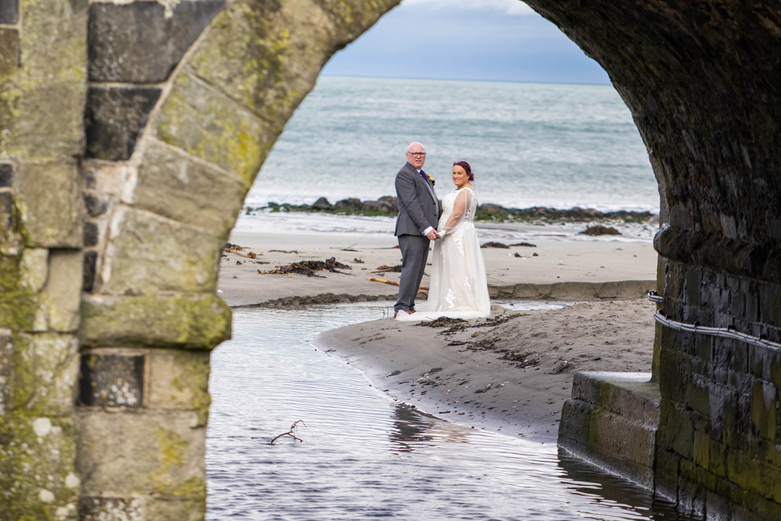 Bride and groom holding hands on a sandy beach, framed by a stone archway, with the ocean in the background.