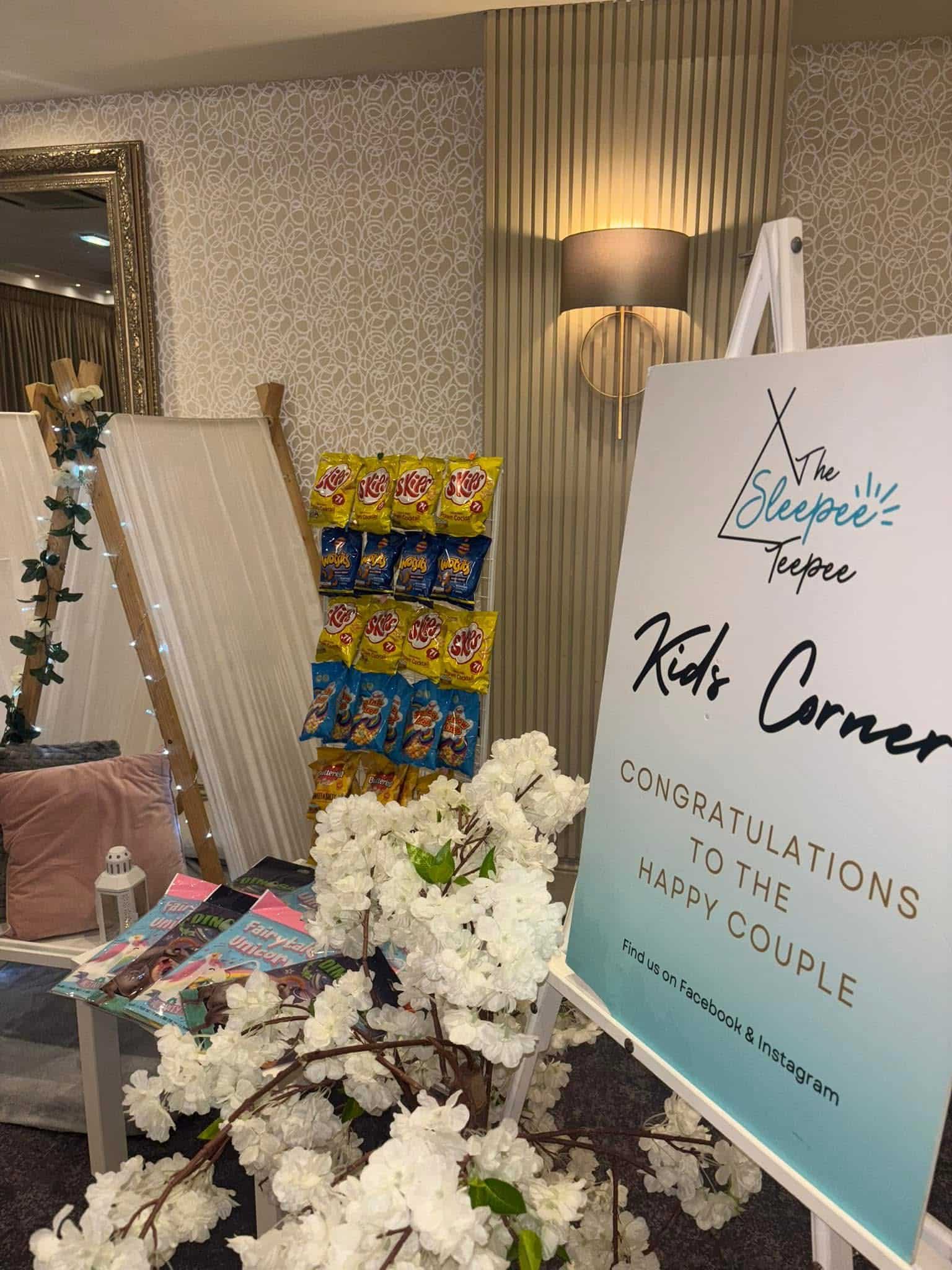 Kids corner setup with snacks, books, and a congratulatory sign for a happy couple, adorned with white flowers.