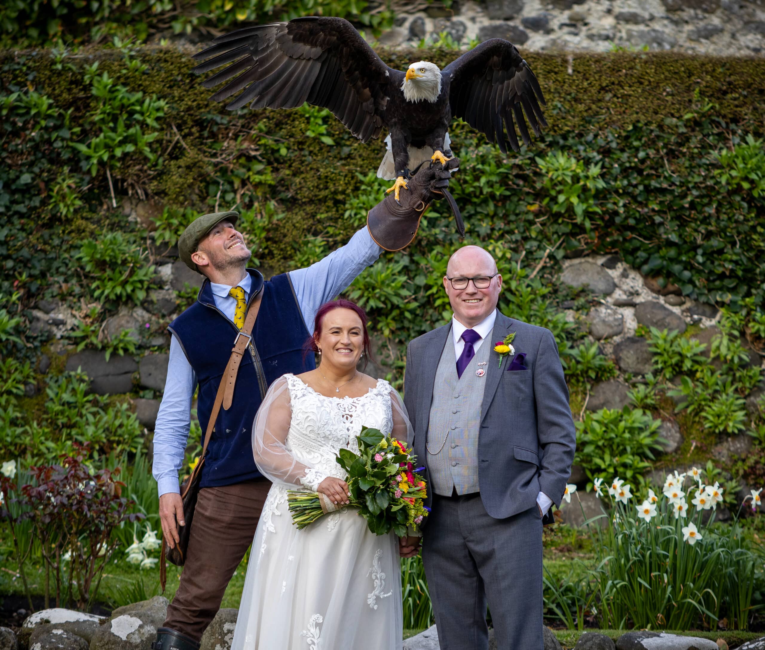 Bride and groom stand with a handler holding a bald eagle, surrounded by lush greenery.