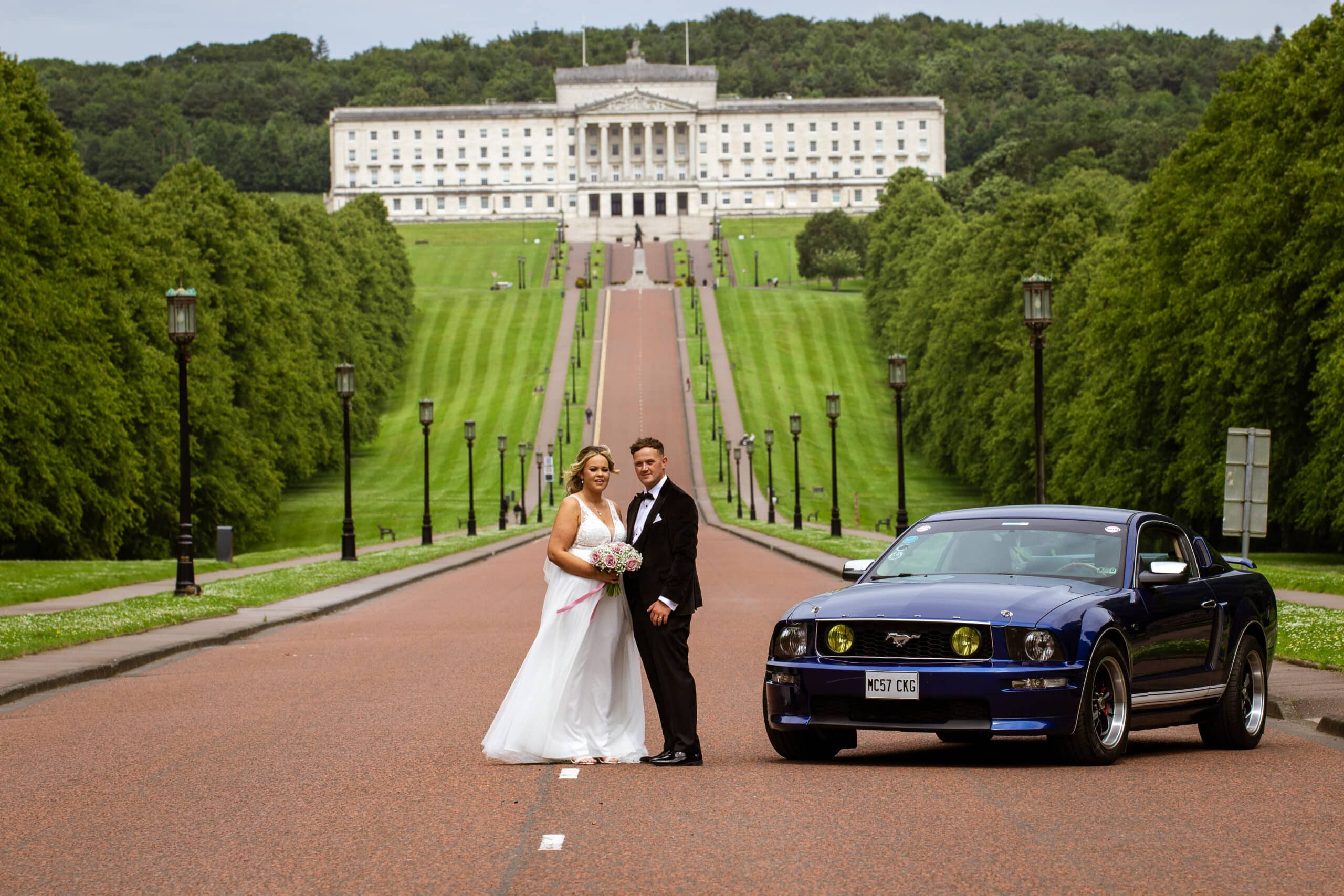 Bride and groom standing on a road with a blue Mustang, large white building in background, lined by green trees.