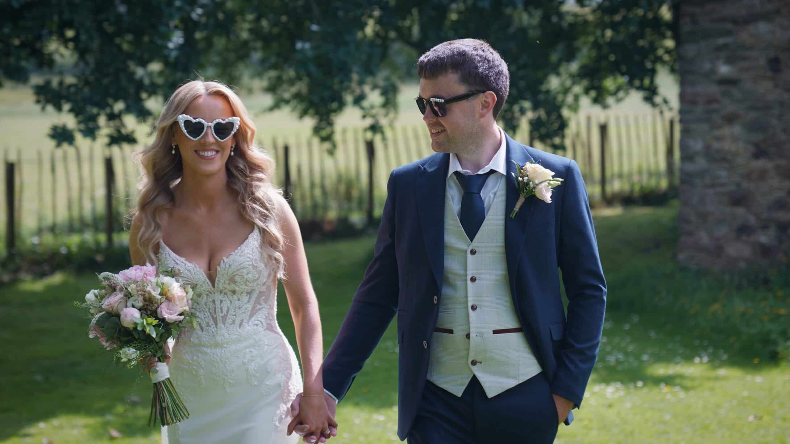 Bride and groom smiling in heart-shaped sunglasses, holding hands, walking outdoors.