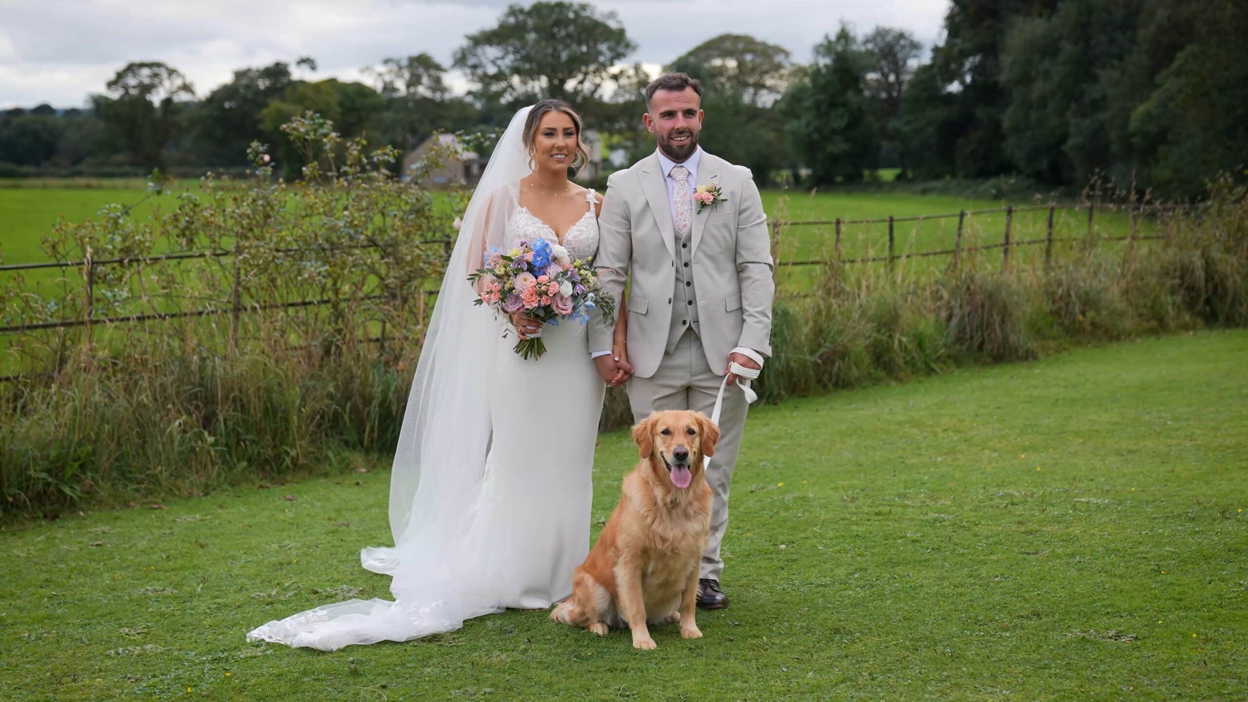 Bride and groom holding hands with a sitting Golden Retriever on grassy field.