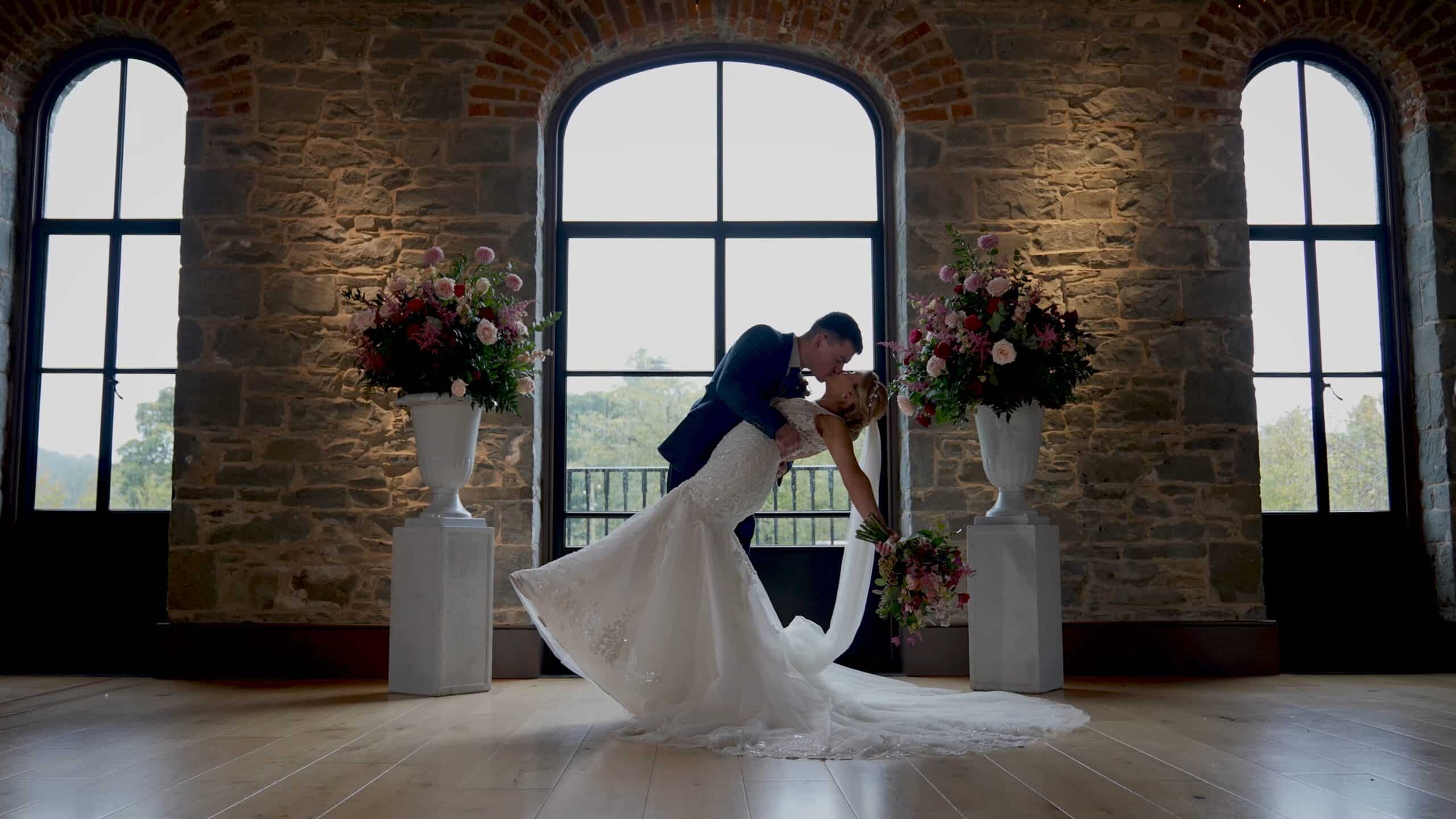 Bride and groom kissing in front of large window, flanked by stone walls and flower arrangements.