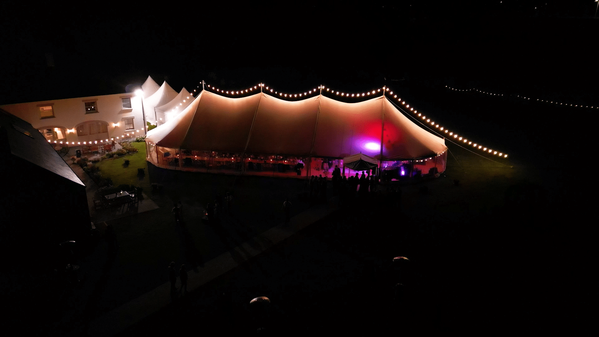 Illuminated marquee tent with string lights at night, set beside a building.