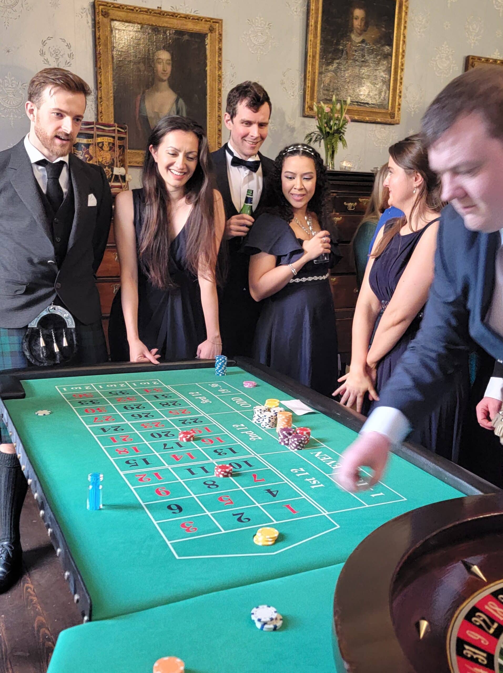 Group of well-dressed people gathered around a roulette table, chatting and watching the game in an elegant room.