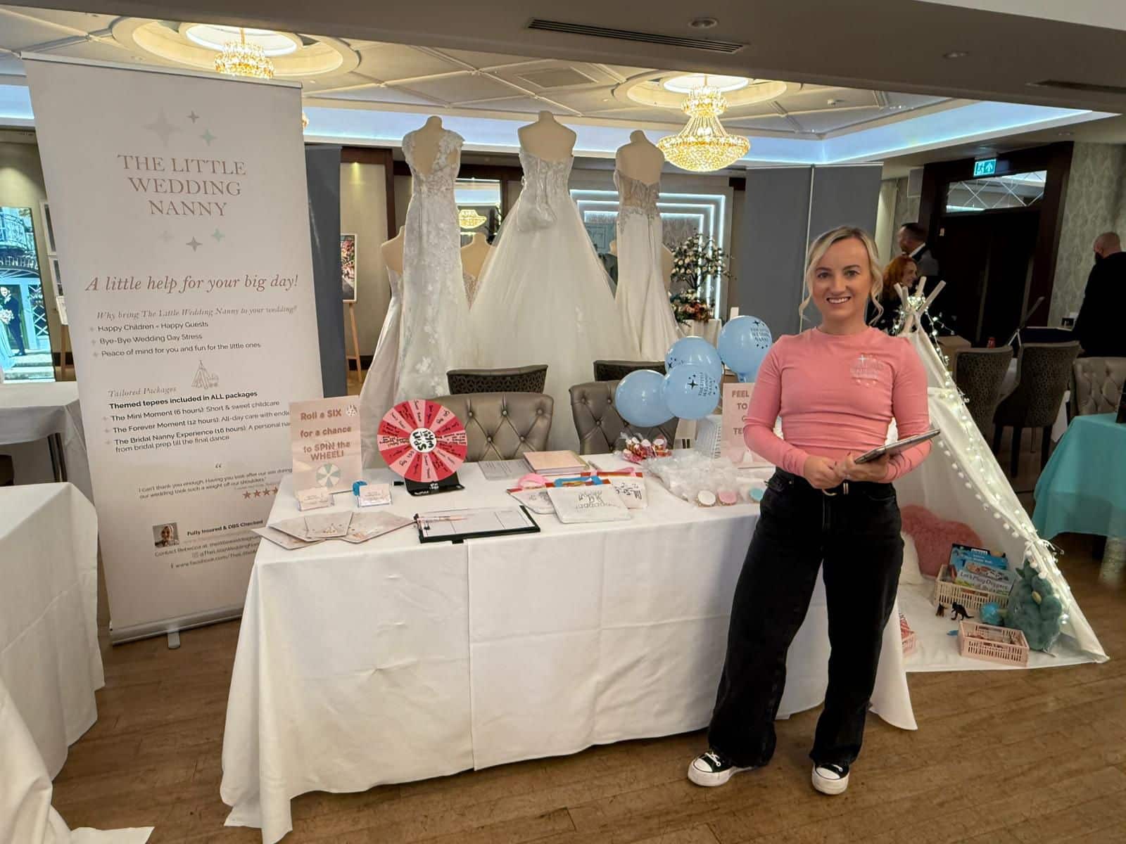 Wedding event booth showcasing gowns and services, with a smiling woman holding a tablet in front.