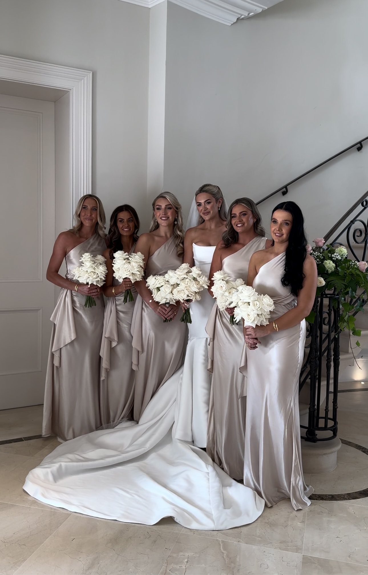 Bride with bridesmaids in elegant dresses holding white bouquets, standing by a staircase.