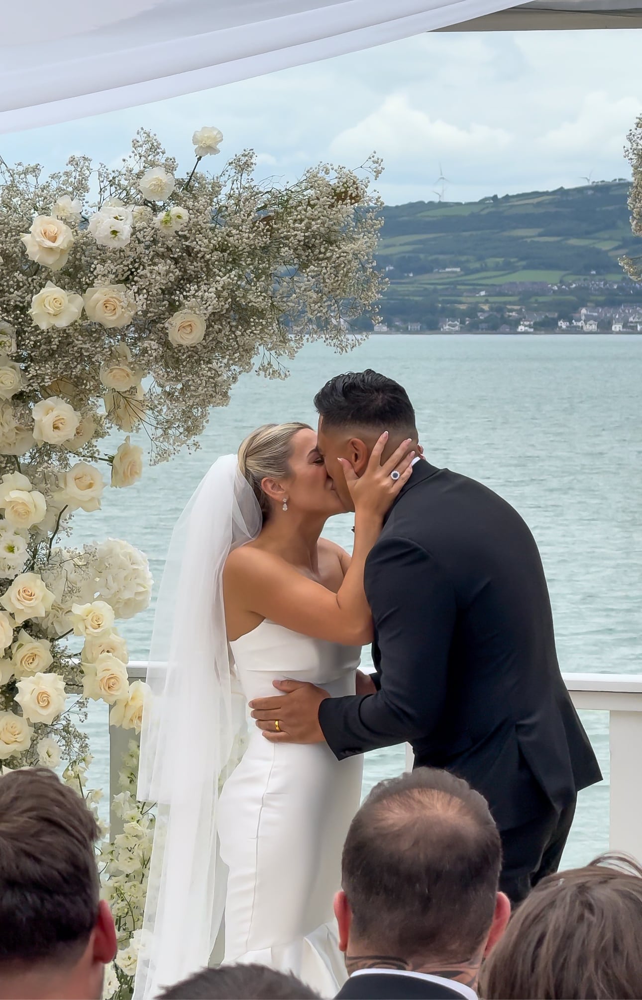Bride and groom kissing at waterfront wedding ceremony, surrounded by white floral arrangements.