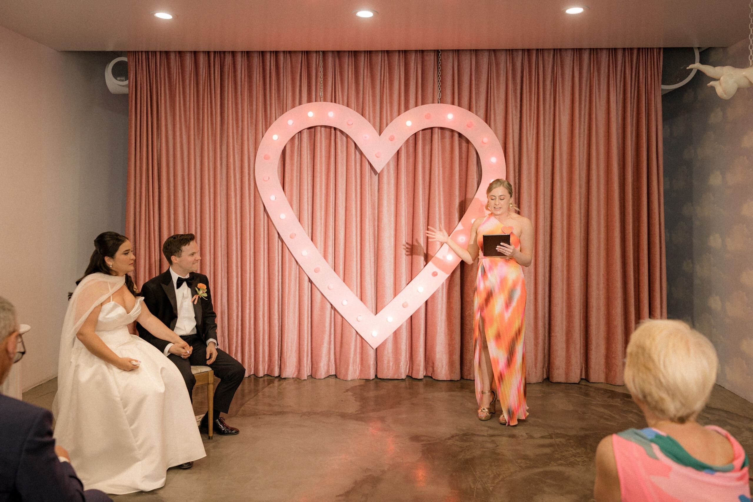 Bride and groom sitting at a wedding ceremony with a large illuminated heart in the background.