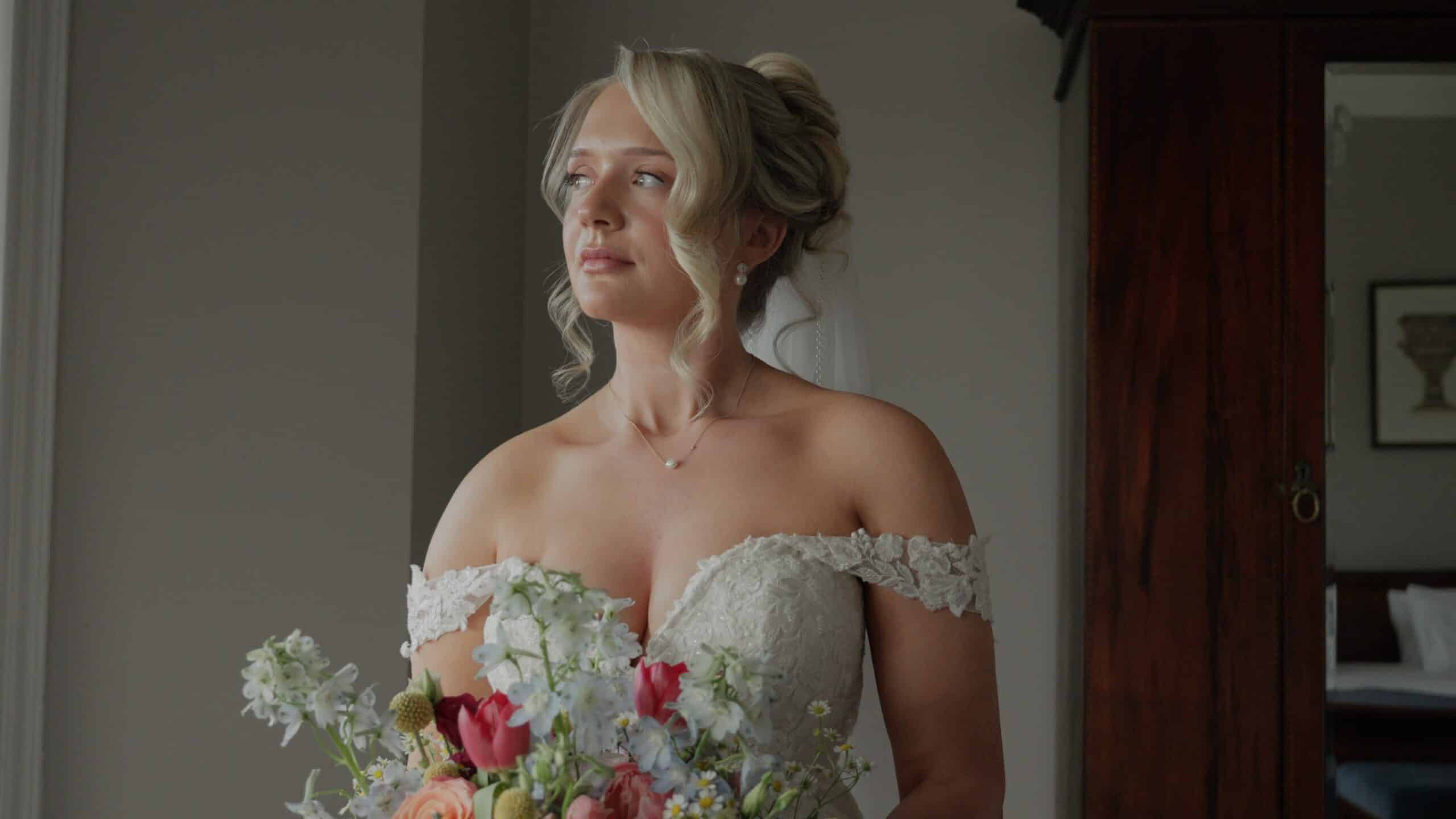 Bride in elegant off-shoulder lace dress holding colorful bouquet, gazing thoughtfully to the side.