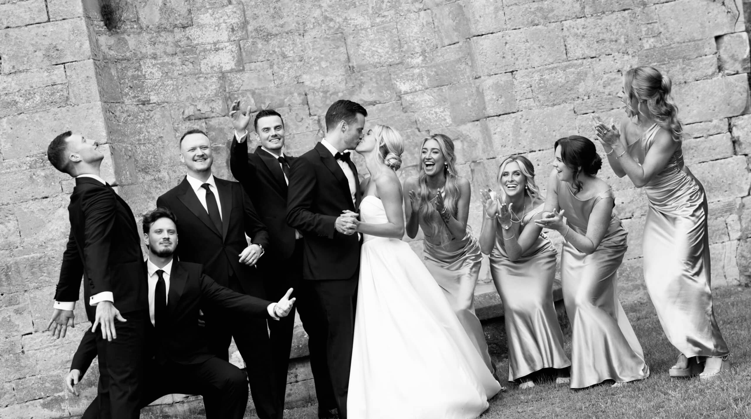 Wedding party cheering as bride and groom share a kiss in front of a stone wall, in black and white.