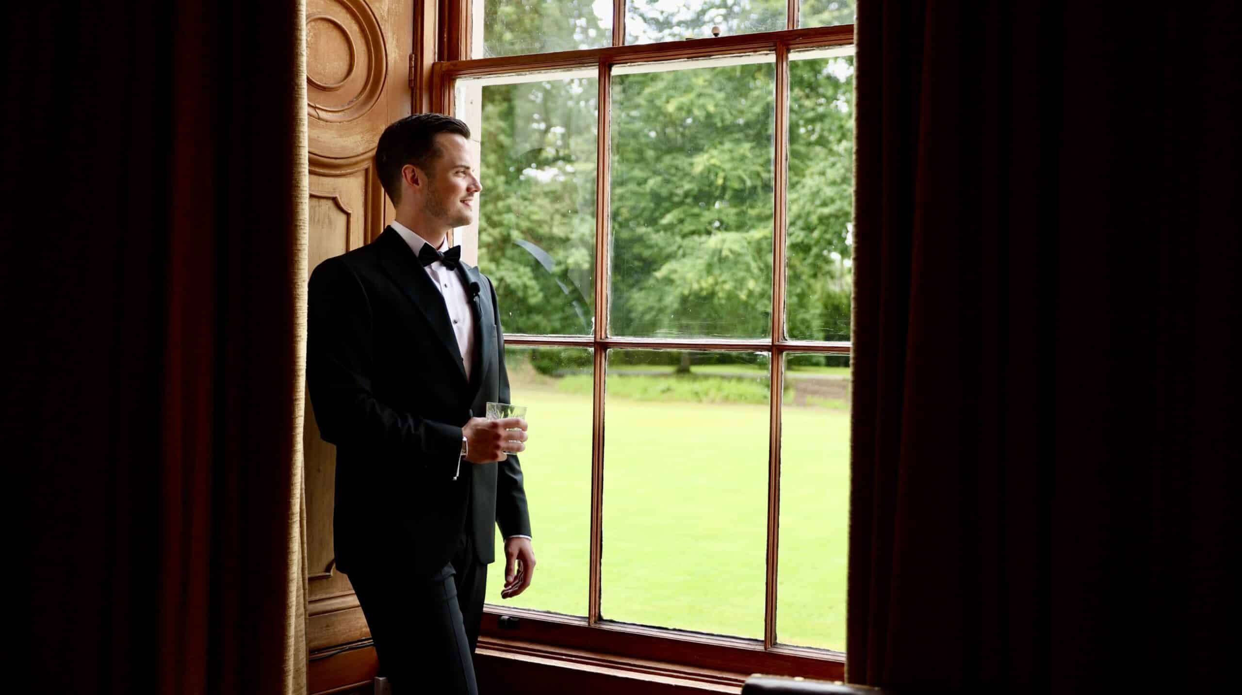 Man in a tuxedo holding a glass, looking out a window at greenery.