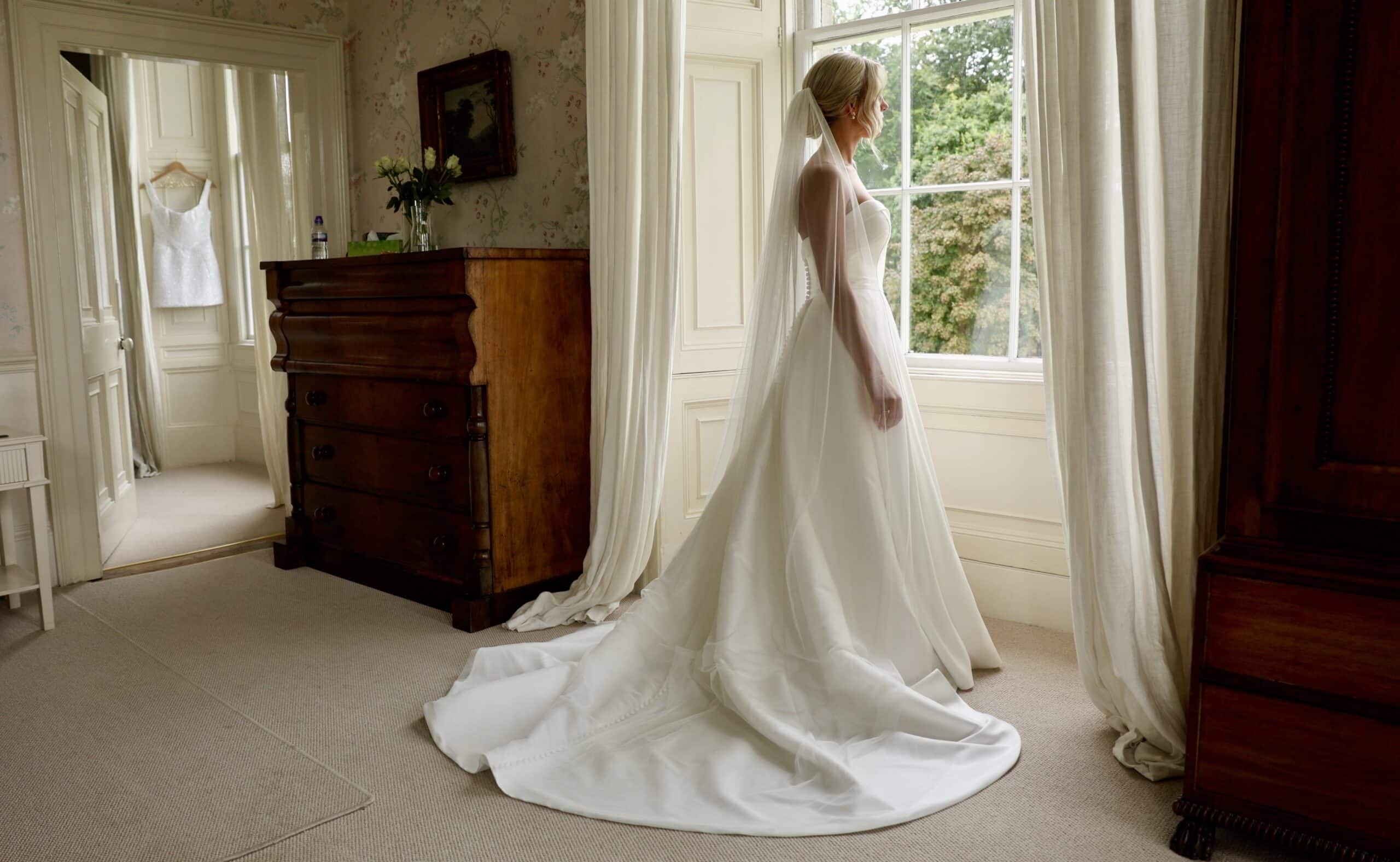 Bride in a white gown looking out a window in a vintage room.