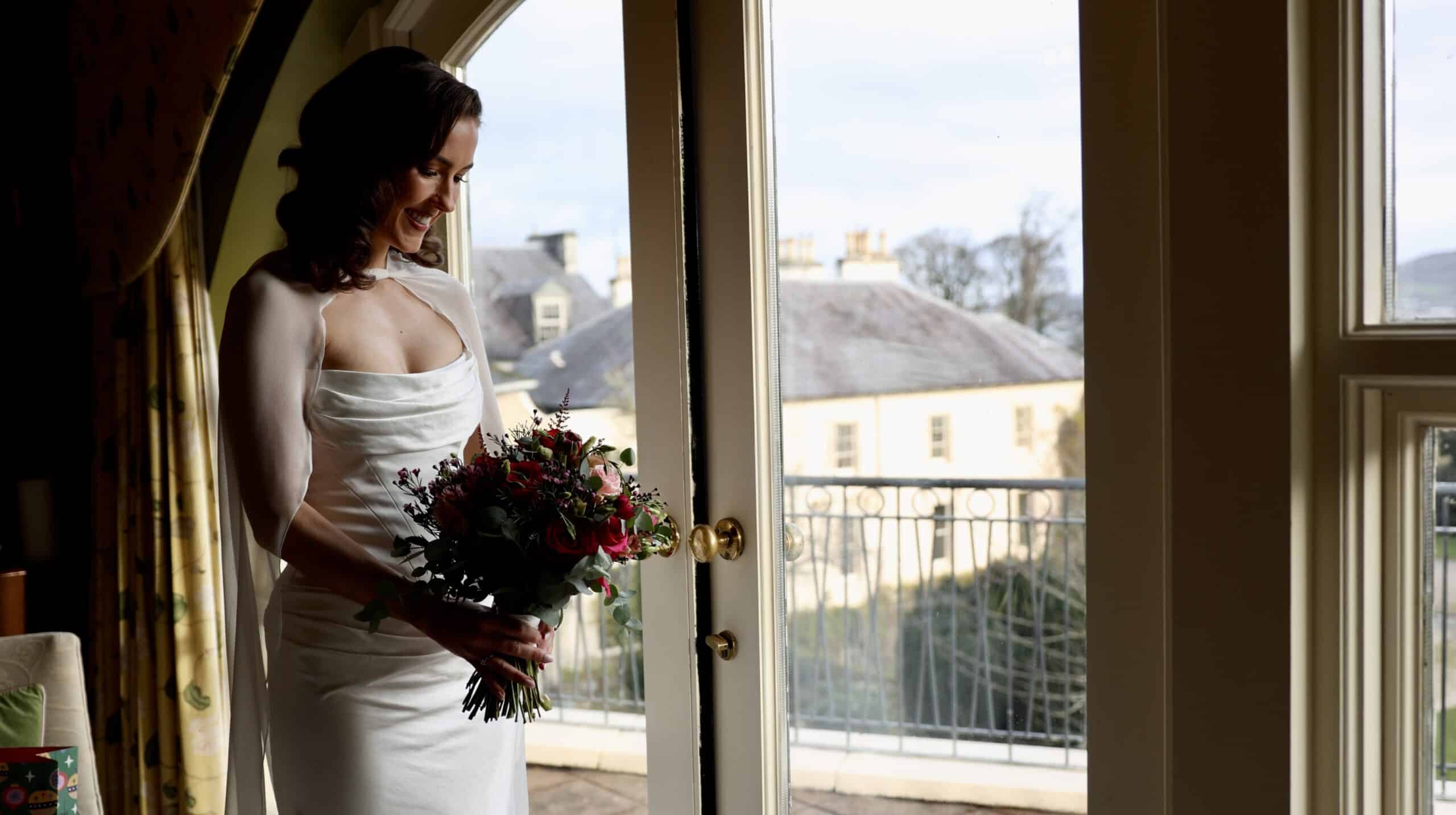 Bride in a white dress holding a bouquet, standing by a window with a scenic outdoor view.