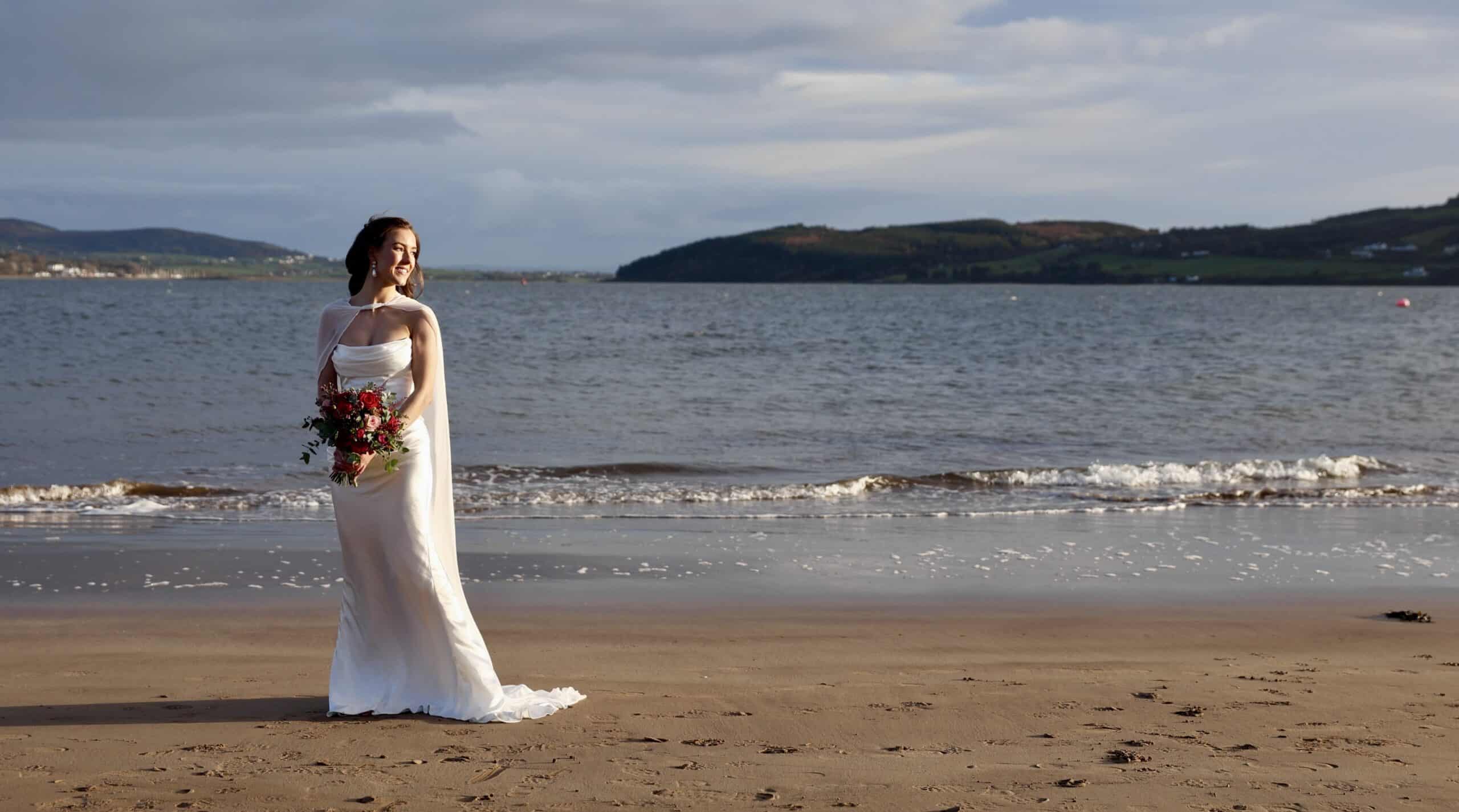 Bride in a white gown holding a bouquet stands on a beach with waves and distant hills behind her.