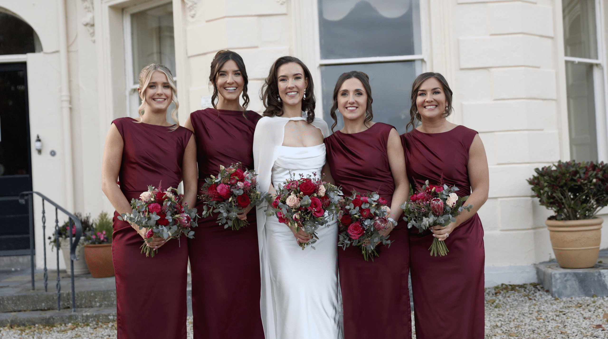 Bride with bridesmaids in burgundy dresses holding bouquets, standing outside a building.