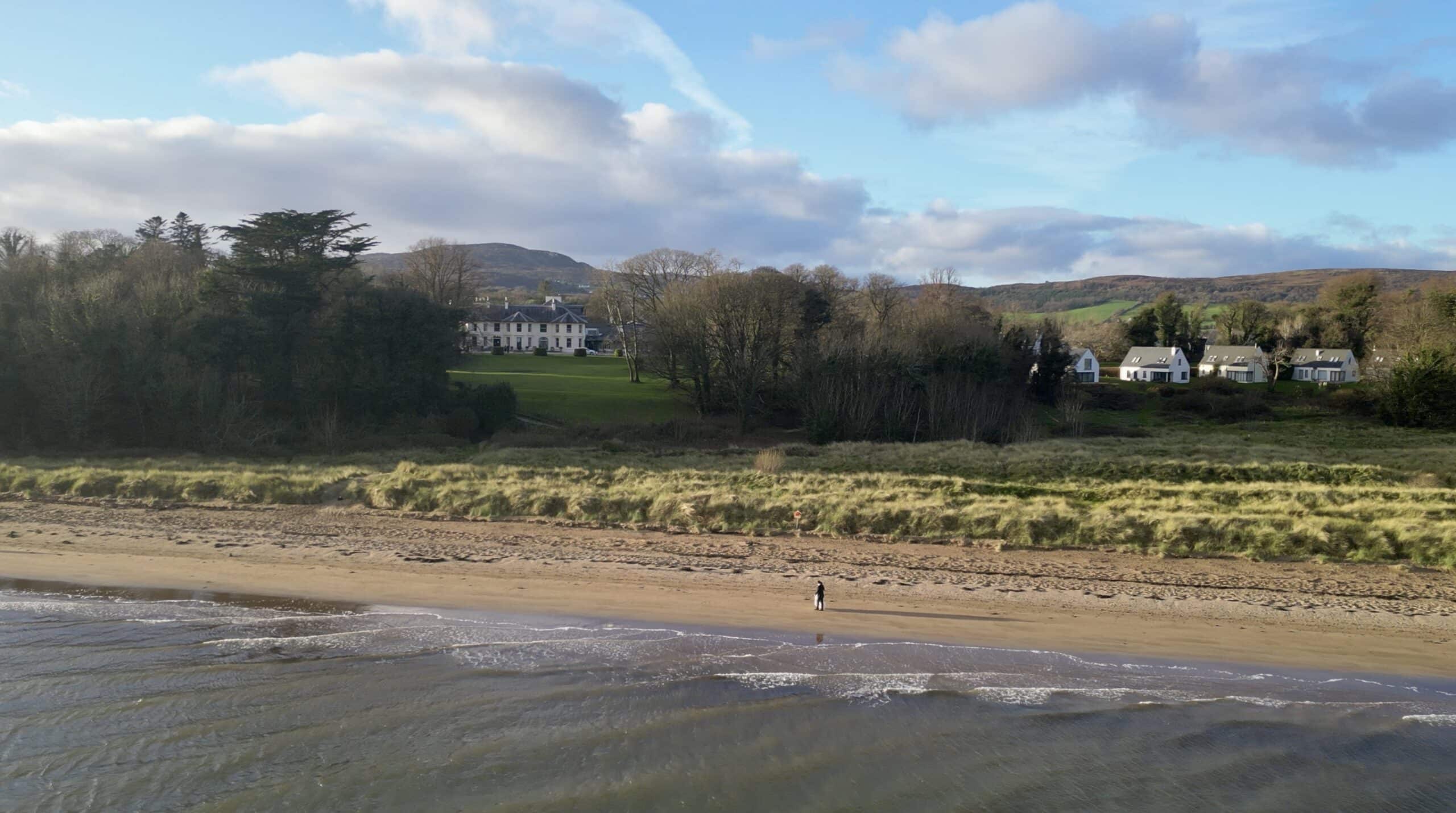 Aerial view of a beach with a single person walking along the shore, surrounded by grassy dunes and houses in the background.
