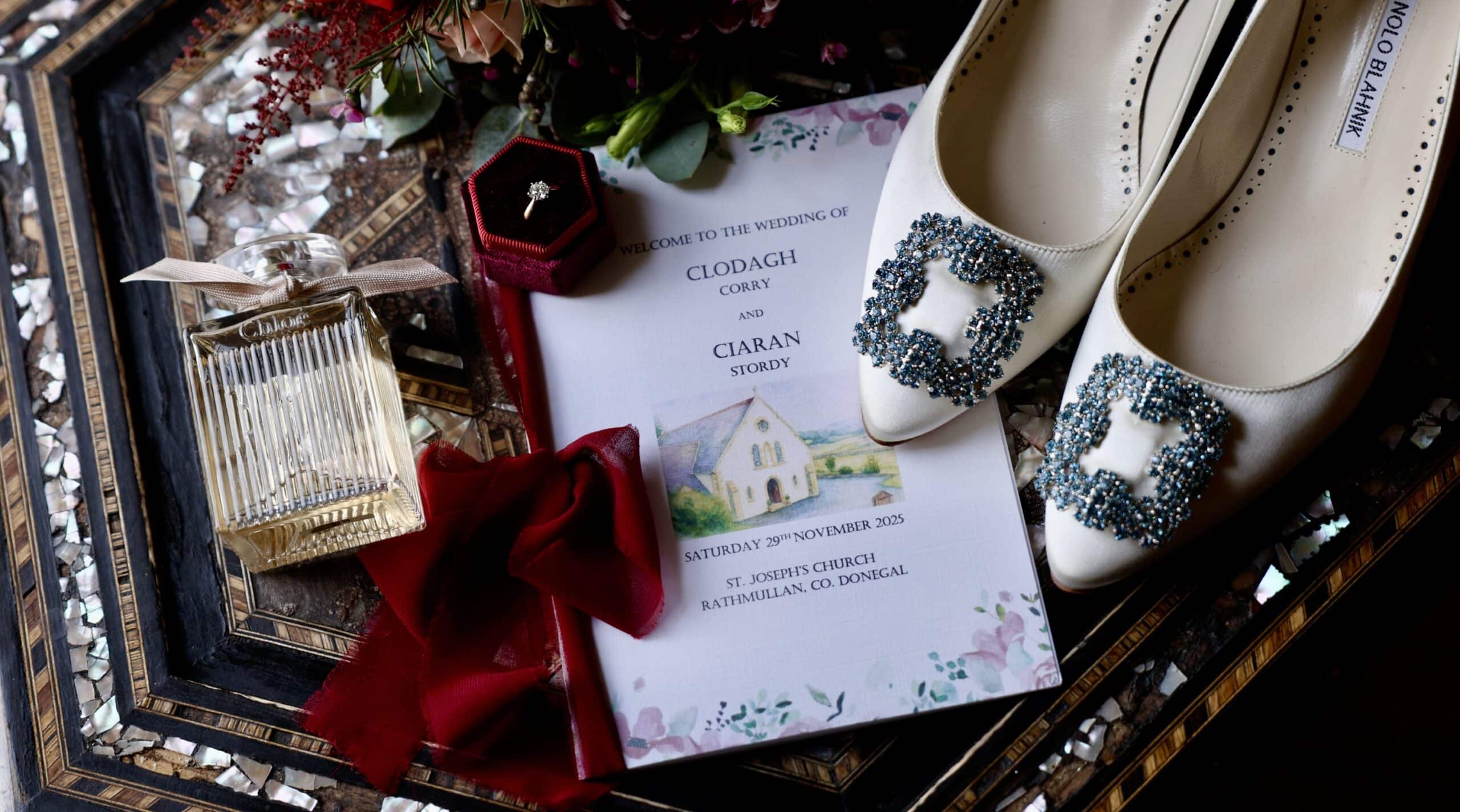 Wedding invitation with shoes, perfume, red ribbon, and a ring box on decorative surface.
