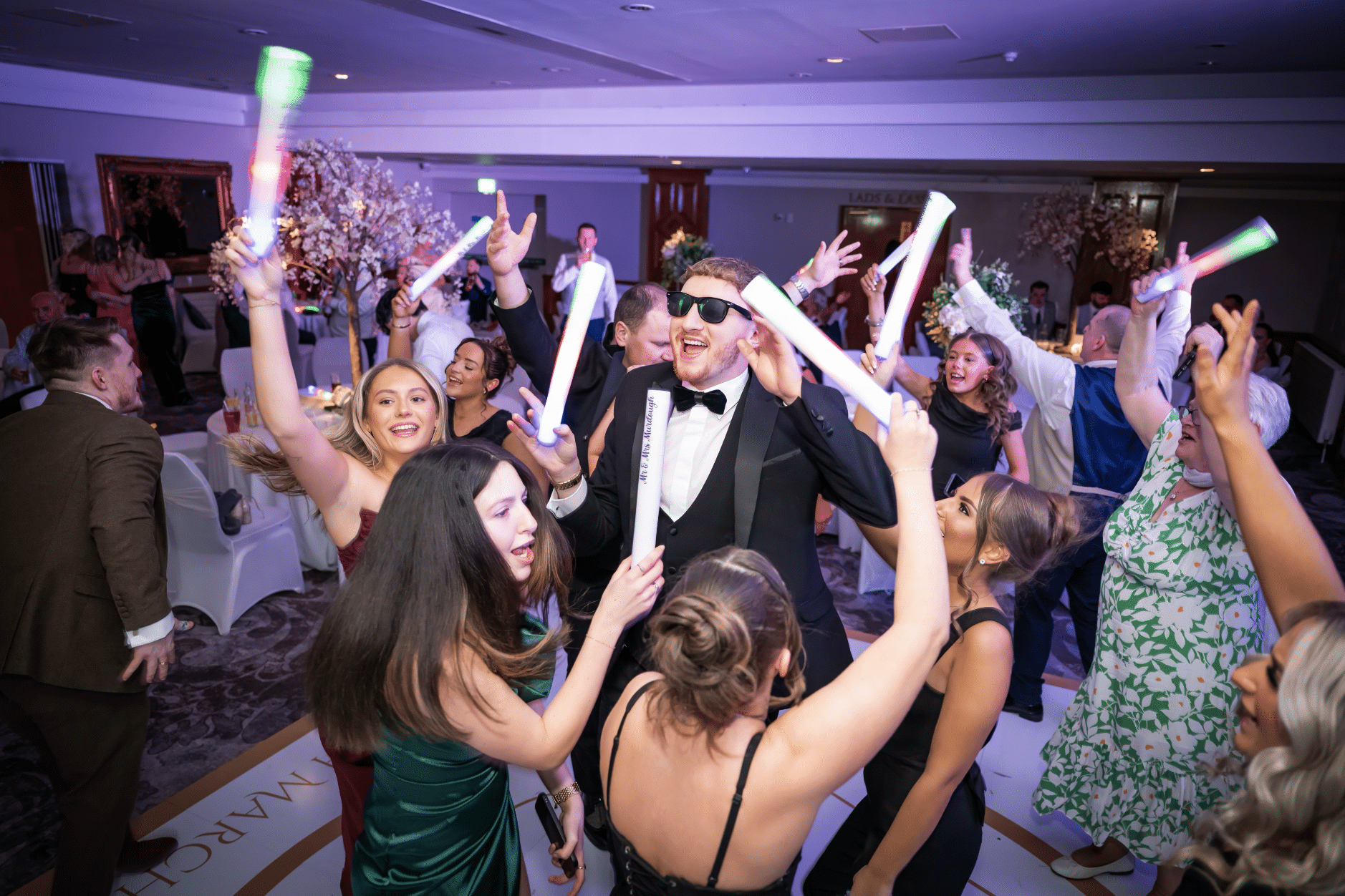 People dancing and celebrating at a party with light sticks in a decorated venue.