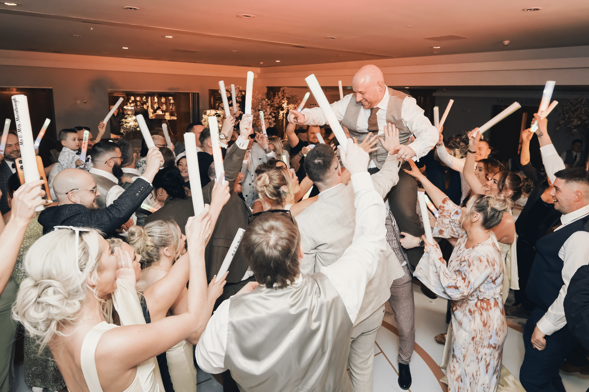 Lively wedding celebration with a group of people raising glow sticks and a man being lifted in the center.