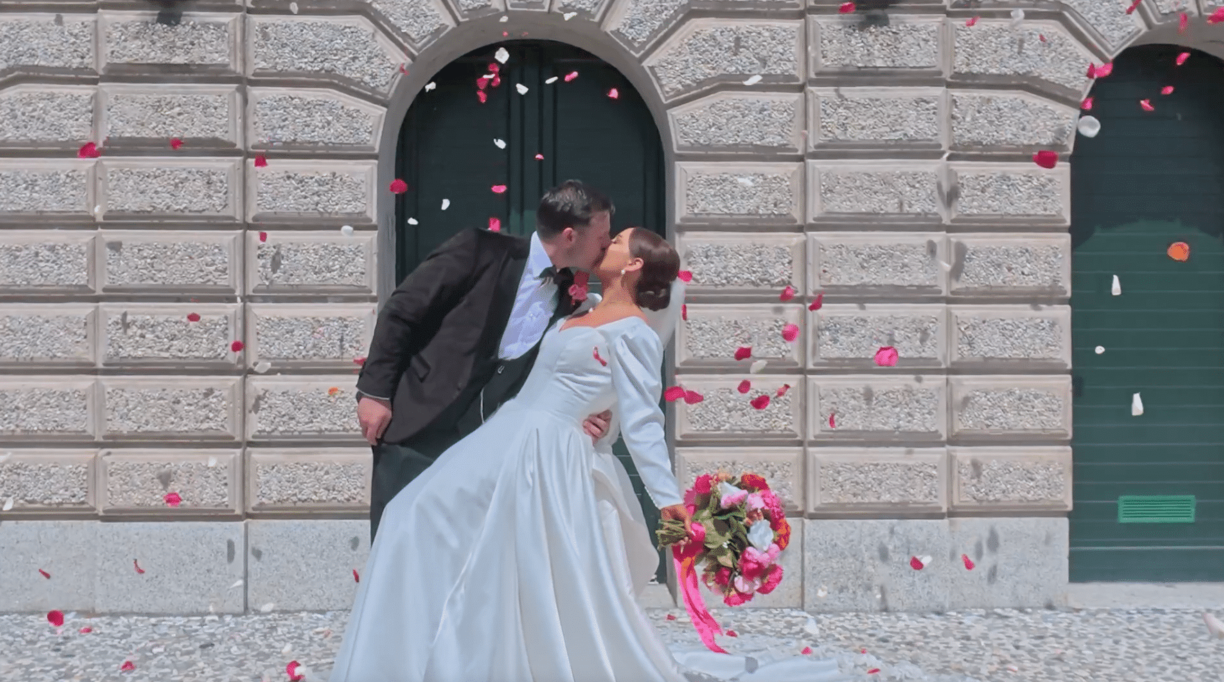 Bride and groom kiss under falling rose petals in front of an arched doorway.