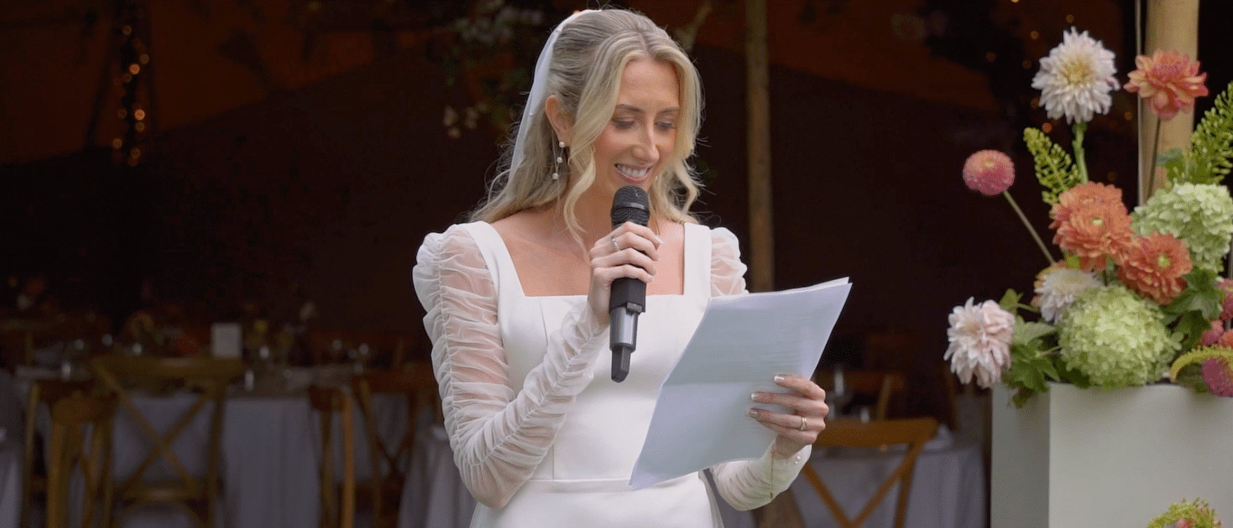 A woman in a white dress gives a speech with a microphone, standing near a floral arrangement.