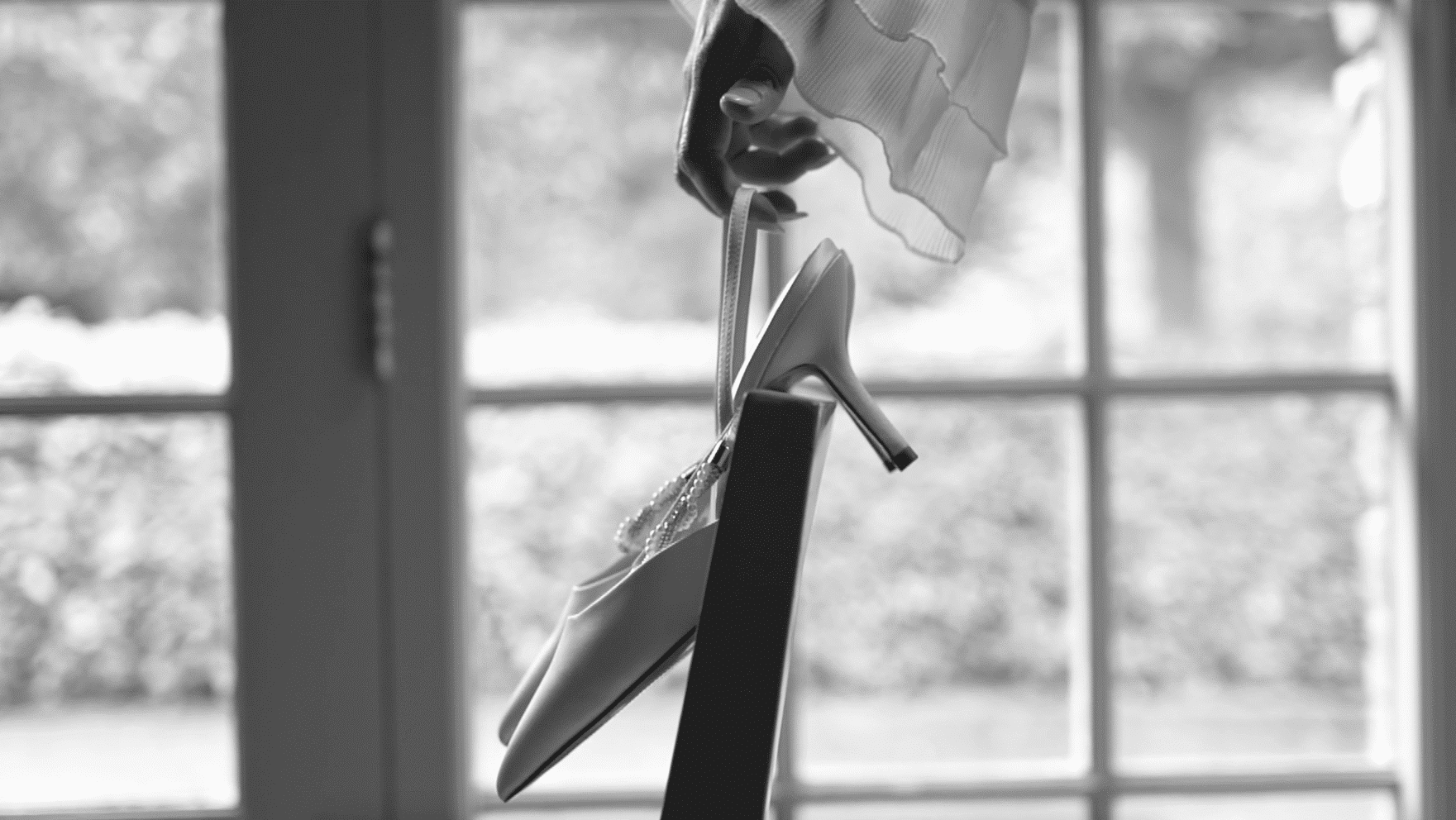 Black and white image of a hand holding a high-heeled shoe beside a window.
