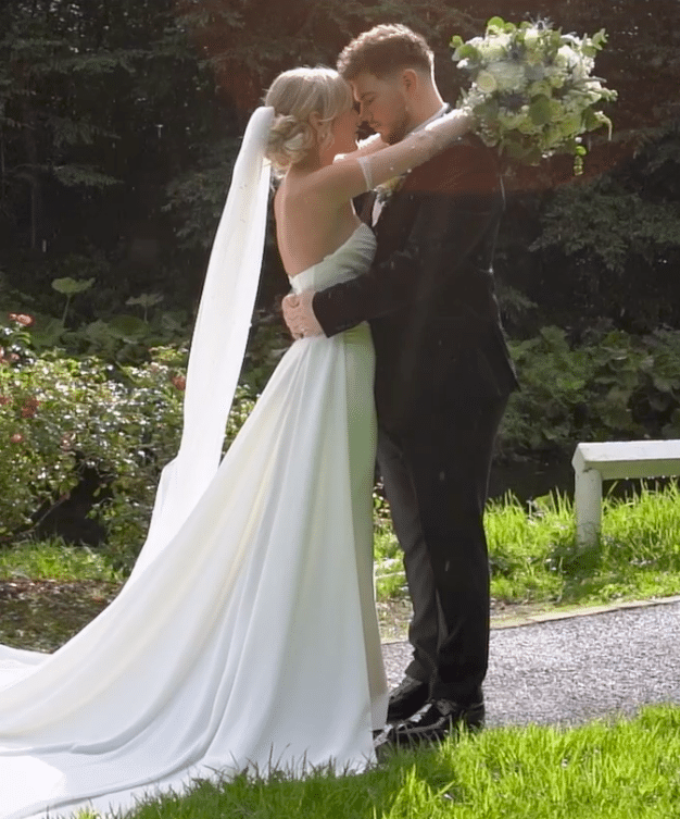 Bride and groom embracing outdoors, bride holding bouquet, with lush greenery in the background.