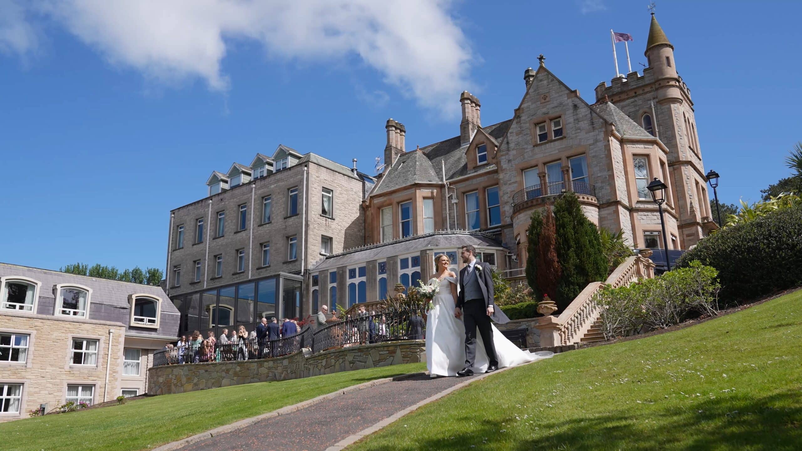 Bride and groom walking down a path, elegant mansion in the background, clear blue sky.