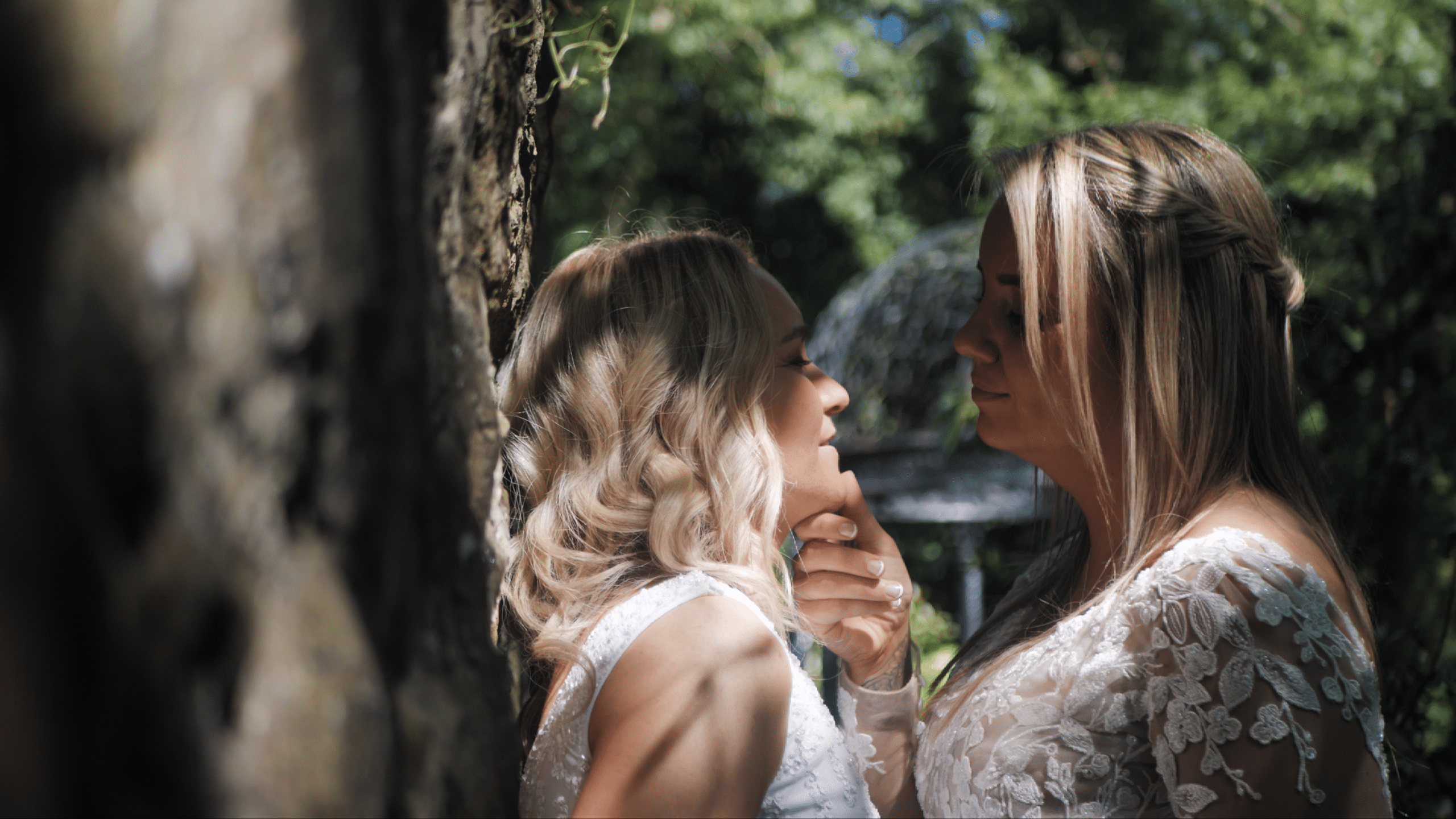 Two women in wedding dresses share an intimate moment outdoors by a tree.