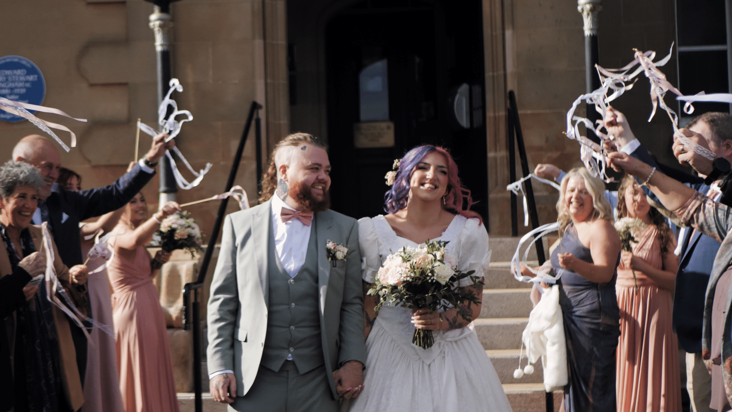 Bride and groom exiting a building, smiling, surrounded by guests waving ribbons, with the bride holding a bouquet.