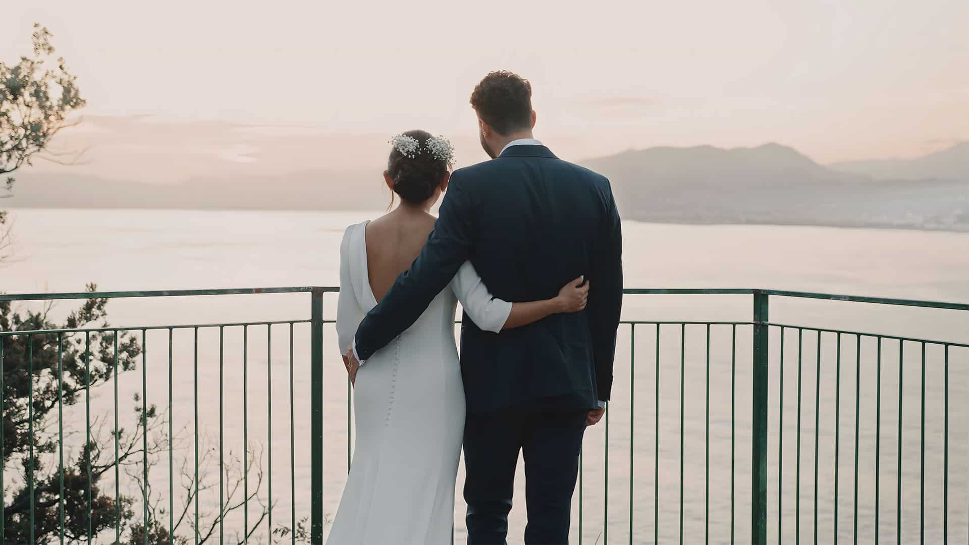 Bride and groom embracing, overlooking a serene sunset by the water.