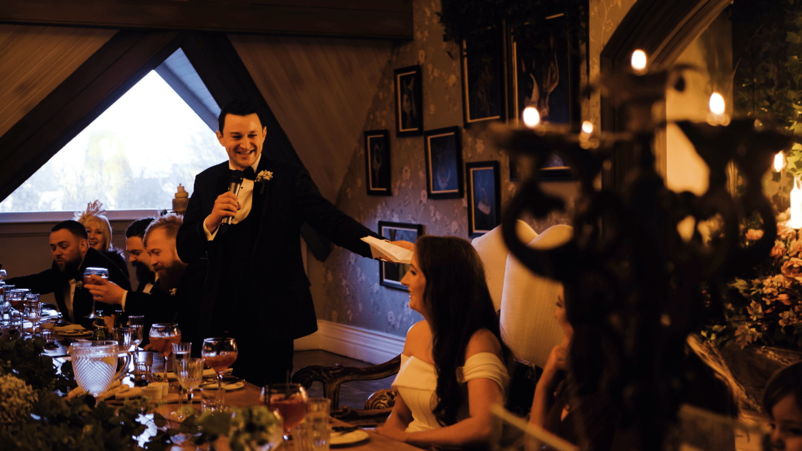 Wedding toast at reception with smiling man holding microphone and seated guests, dimly lit ambiance with candles and decorations.